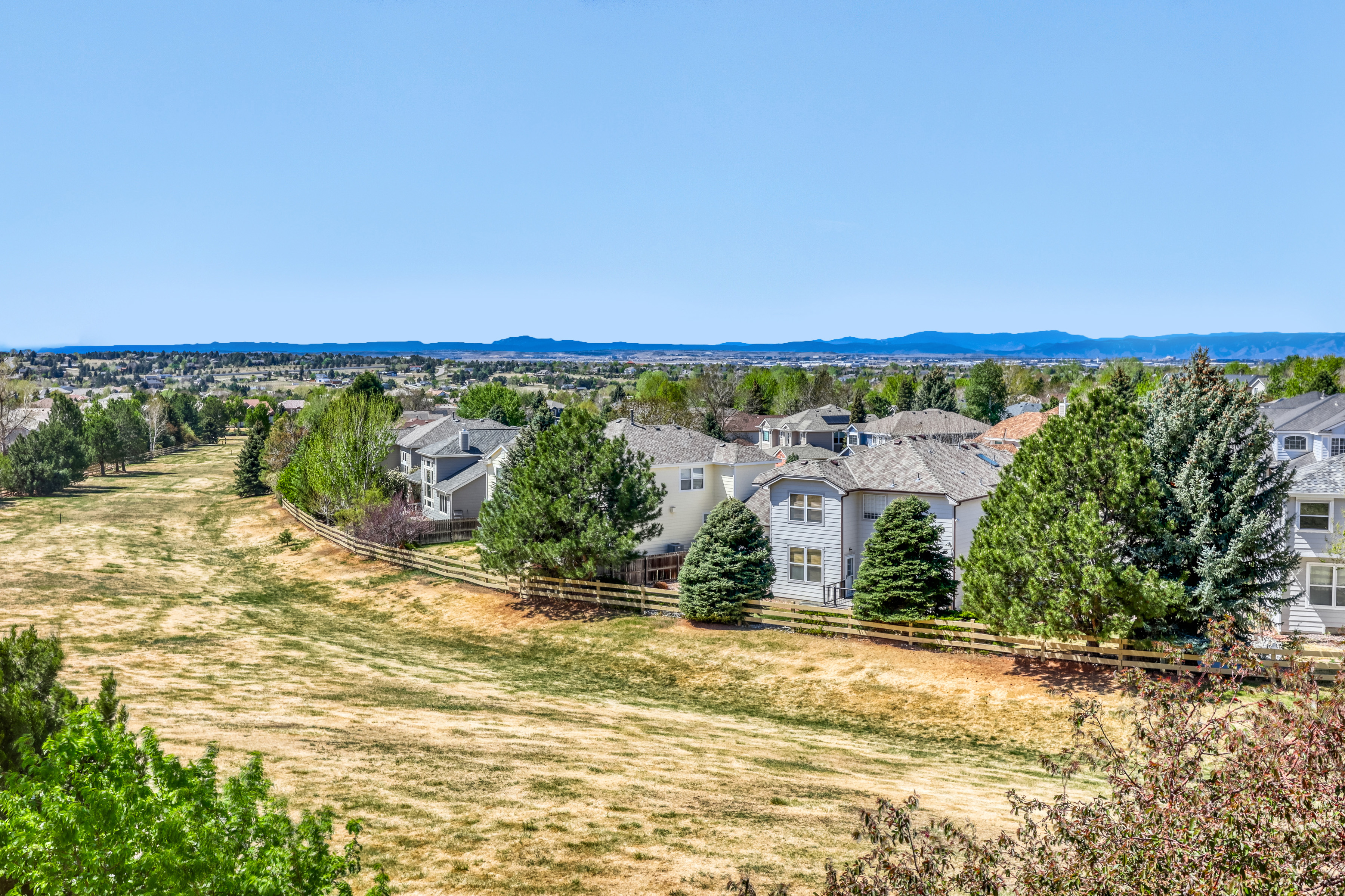This high-angle, aerial perspective showcases a suburban residential neighborhood bordering a large, open grassy field. The scene captures a series of multi-story homes with neutral siding and peaked roofs, separated from the common area by a decorative wooden fence. The setting offers a peaceful and spacious atmosphere with mature trees and a clear view toward distant rolling hills.