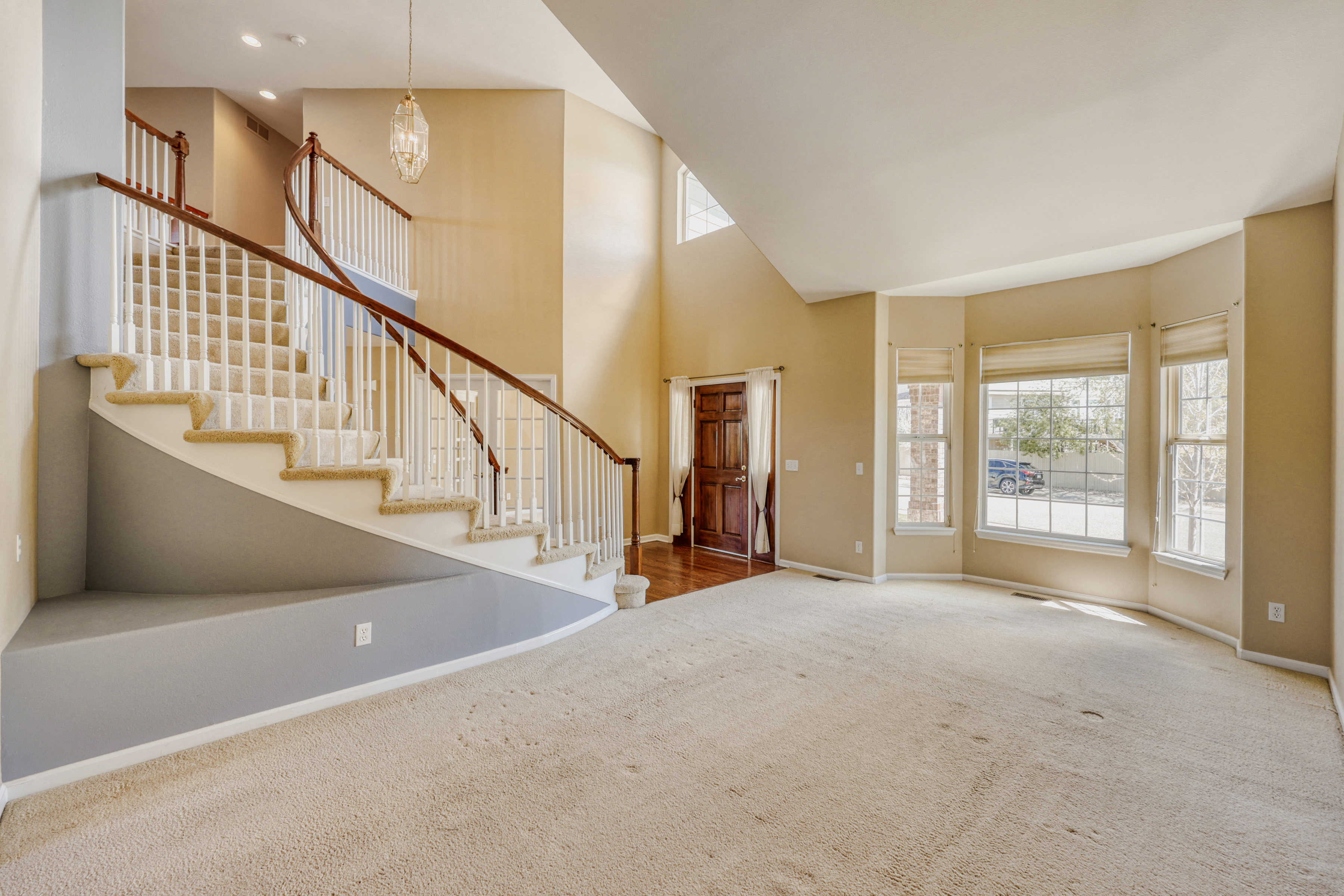 This spacious foyer features a grand, curved staircase with beige carpeting and a polished wood handrail. The open-concept design is enhanced by high ceilings, neutral beige walls, and natural light streaming in through a large bay window and a decorative front door. This area serves as a formal entry and transition point, highlighted by a contemporary tiered chandelier.