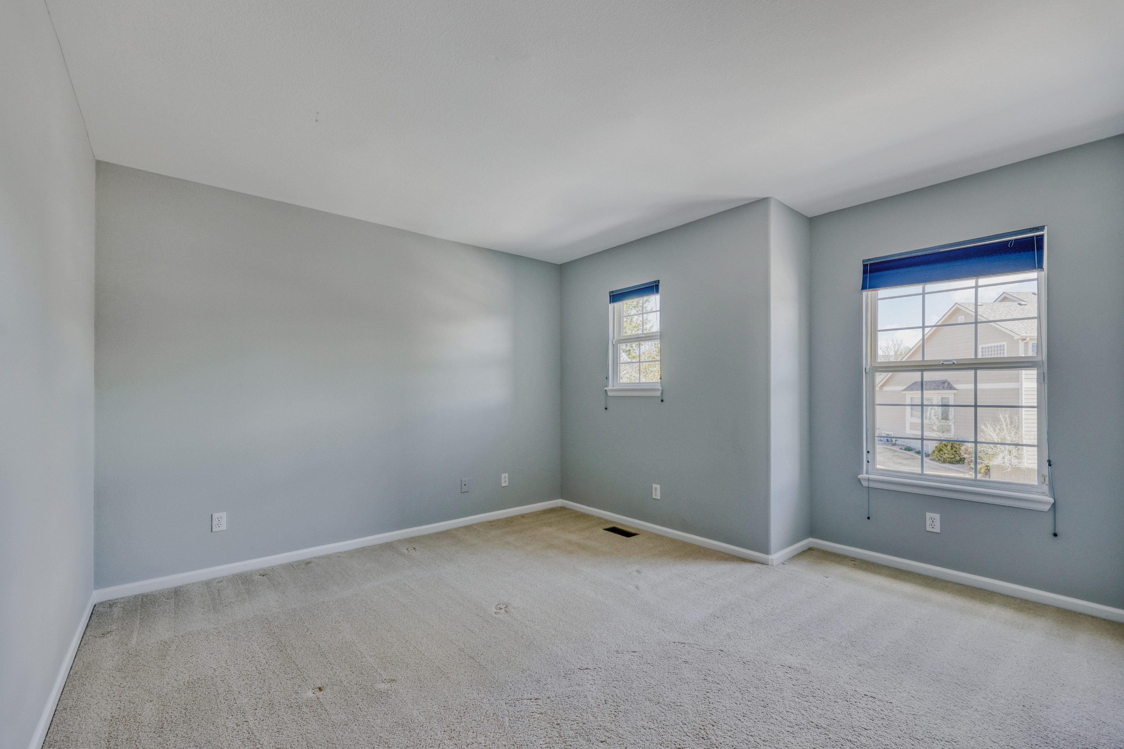 This is a quiet, empty bedroom featuring light grey walls and beige carpeting. Two windows with blue blinds provide natural light and views of the neighborhood. The room offers a clean canvas, making it ideal for use as a guest bedroom, home office, or playroom.