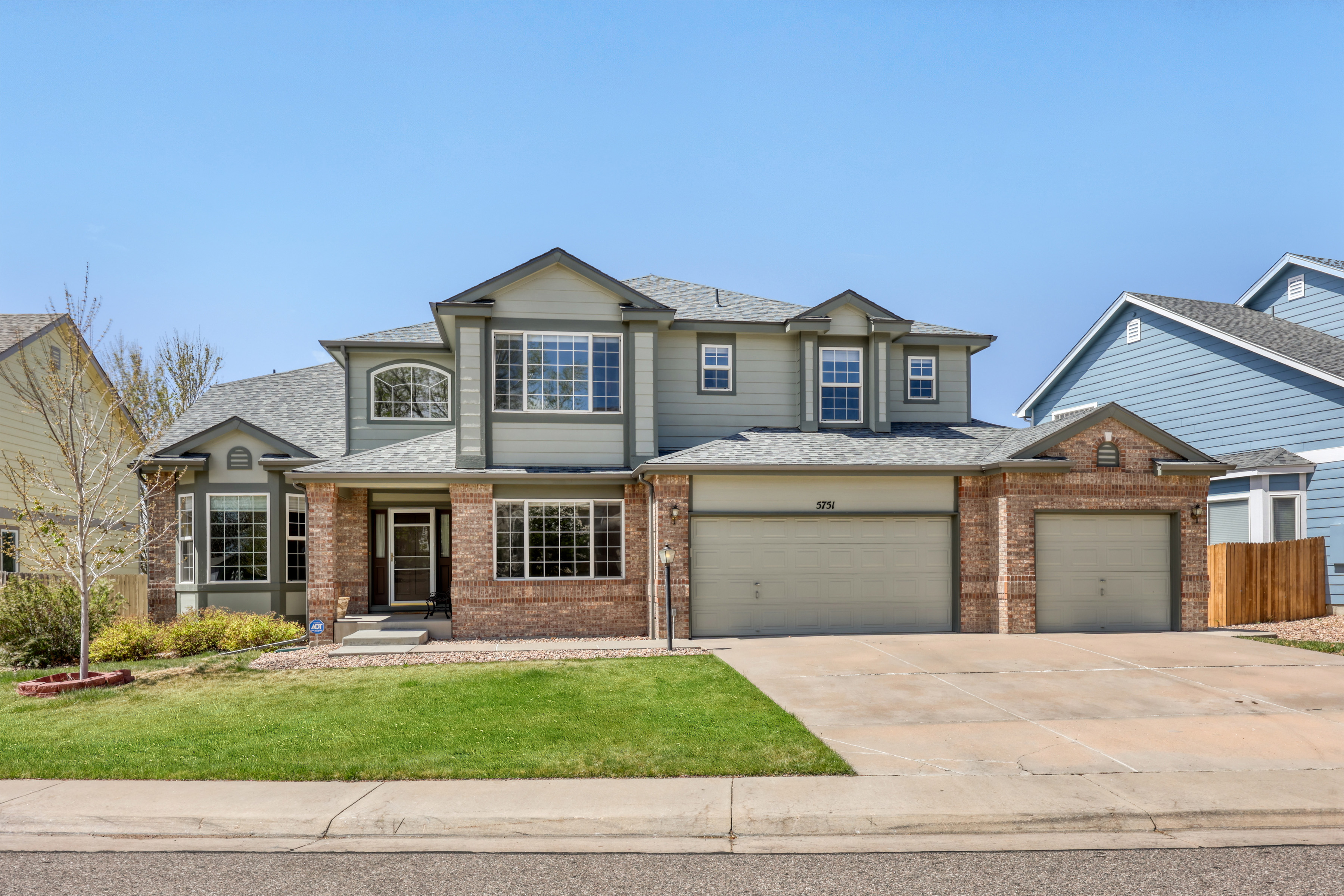 This two-story suburban home features a traditional blend of brick and light green siding with a well-maintained front yard. It includes an attached three-car garage, a covered entryway, and a variety of window designs, providing ample curb appeal. The residence is situated on a clean, paved street in a residential neighborhood with clear blue skies.