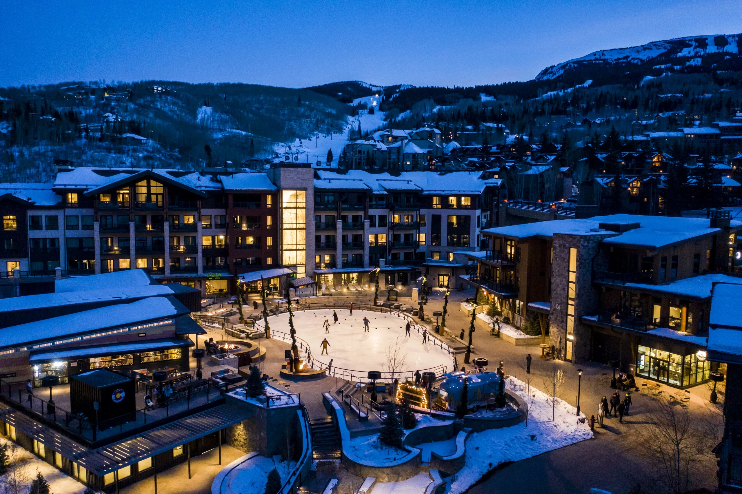 This aerial shot presents a luxurious ski resort community at night. A central ice-skating rink is surrounded by elegantly lit buildings with warm illumination spilling from the windows. Snow-covered rooftops and landscape elements add to the winter ambiance, highlighting the upscale and inviting atmosphere of this real estate offering.
