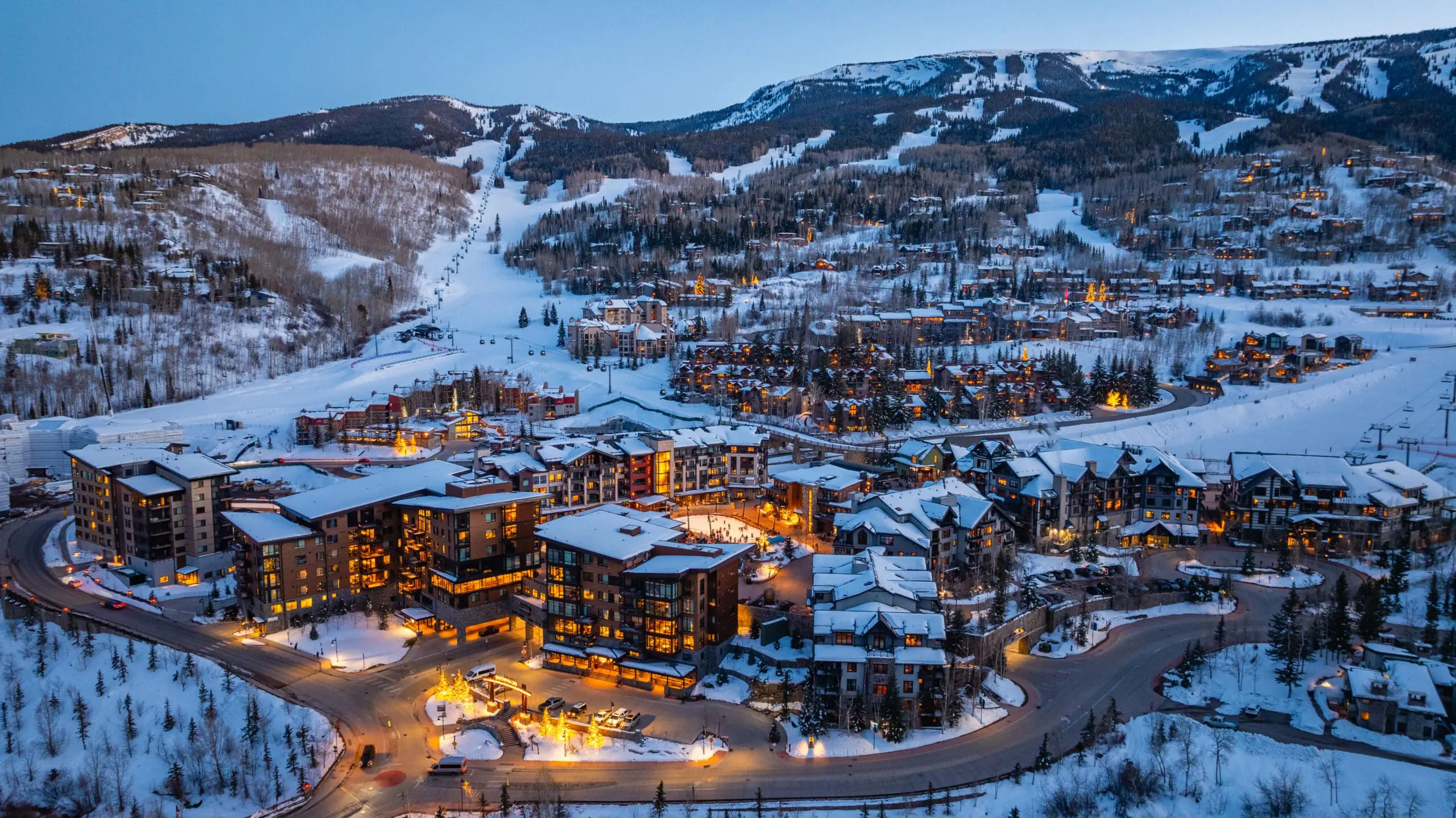This aerial view captures a ski resort town at dusk, showcasing multi-story buildings with snow-covered roofs and warmly lit interiors. Winding roads connect the properties, and snow-laden trees provide a scenic backdrop. The overall impression is luxurious and inviting, highlighting the resort's appeal for winter getaways.