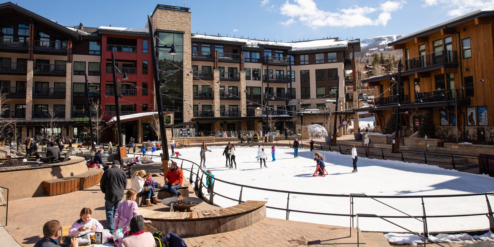 The image shows a lively outdoor community space featuring an ice-skating rink and surrounding seating areas with fire pits. Modern multi-story buildings create a backdrop, offering a sense of upscale living within the community. This amenity promotes a recreational and social lifestyle for residents.