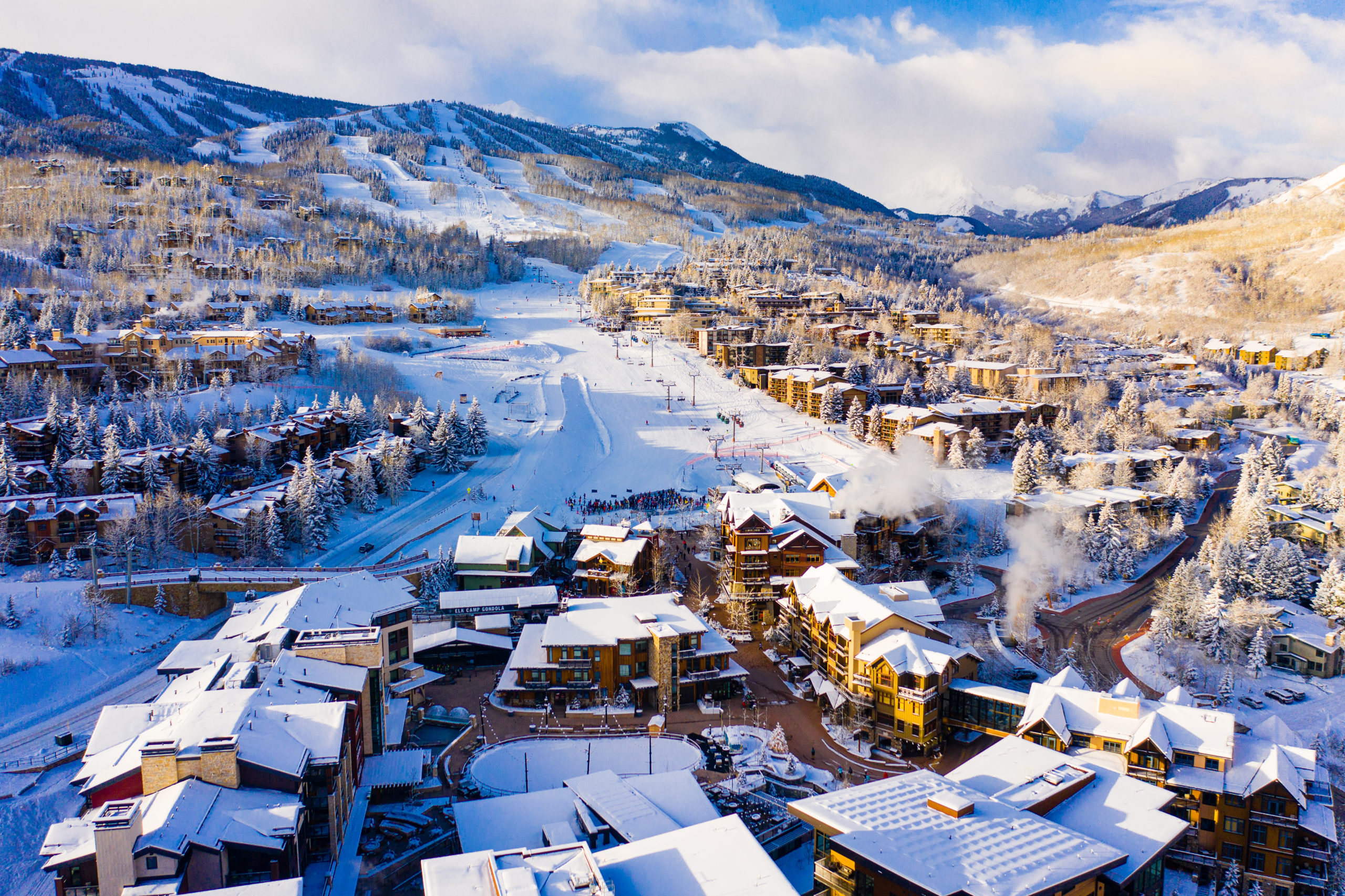 This aerial view showcases a luxurious ski resort community nestled in a snow-covered mountain landscape. The image highlights numerous chalets and buildings with snow-laden roofs, connected by ski runs and roads. The pristine white snow against the architectural elements gives an impression of a premium winter retreat.
