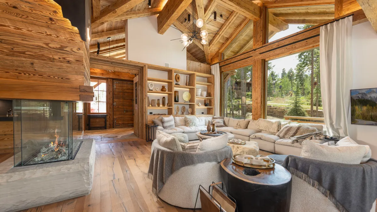 This image showcases a luxurious living room with a rustic, chalet-style design. Key features include exposed wooden beams and wood-paneled walls, a modern glass fireplace, and comfortable, oversized seating arrangements. Large windows offer views of the outdoor landscape, creating a warm and inviting atmosphere.