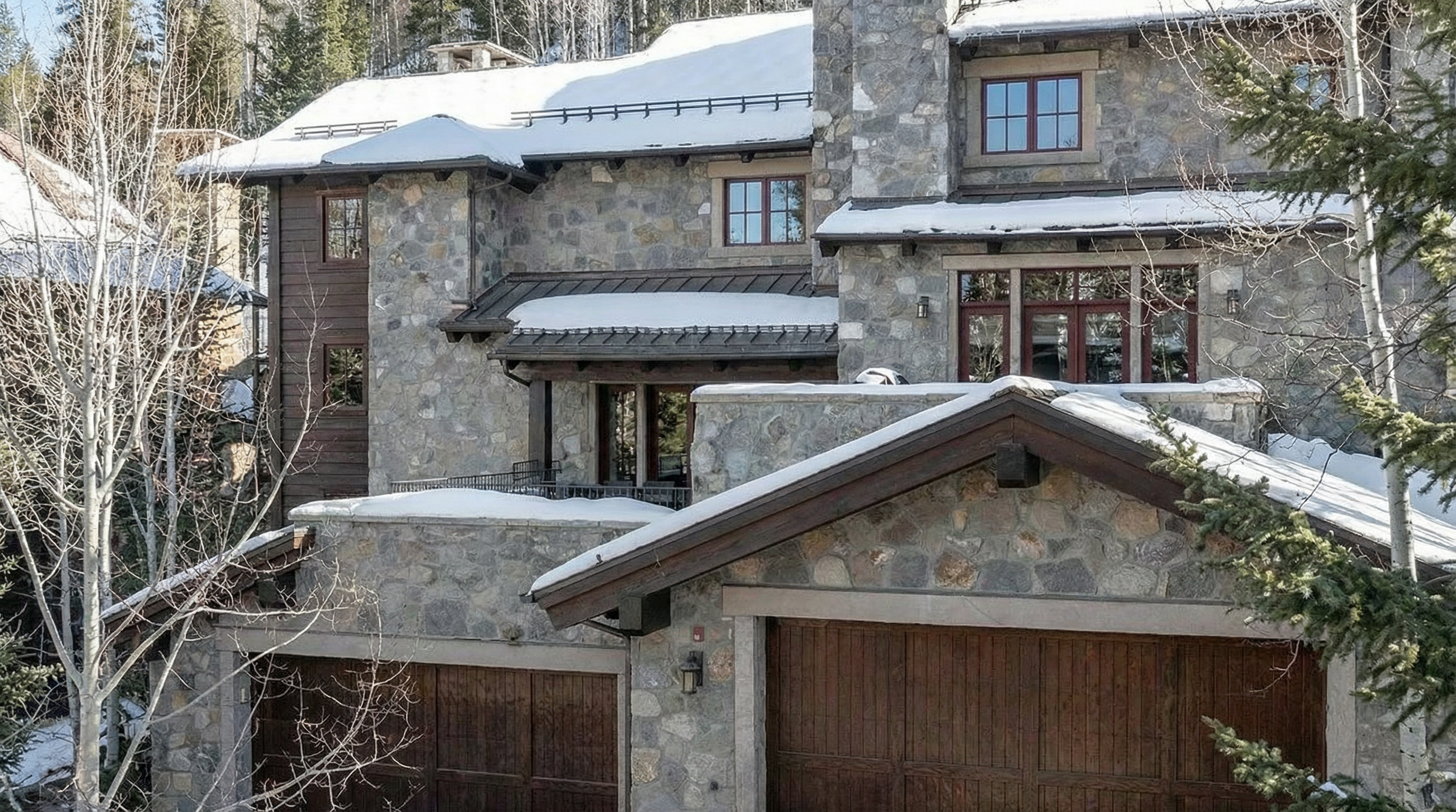 This is a beautiful stone house with a sophisticated design, likely located in a snowy mountain area. The exterior showcases a combination of stone and wood, with multiple levels and visible garage doors. Snow adorns the roof, adding to the picturesque, wintery ambiance of the property.