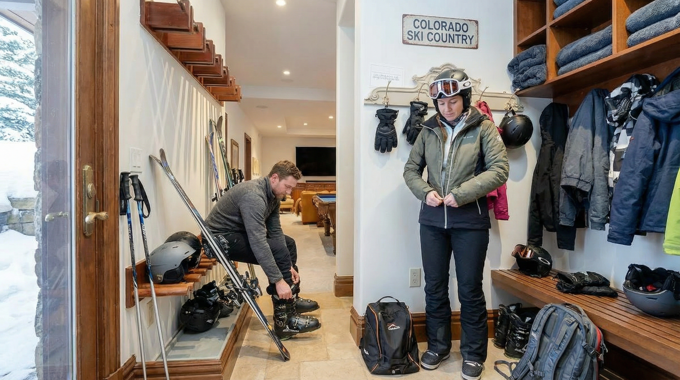 This interior shot captures a ski lodge-style entryway. The scene features a man putting on ski boots, a woman zipping up her jacket, and ski equipment neatly arranged. A "Colorado Ski Country" sign adds to the thematic decor.