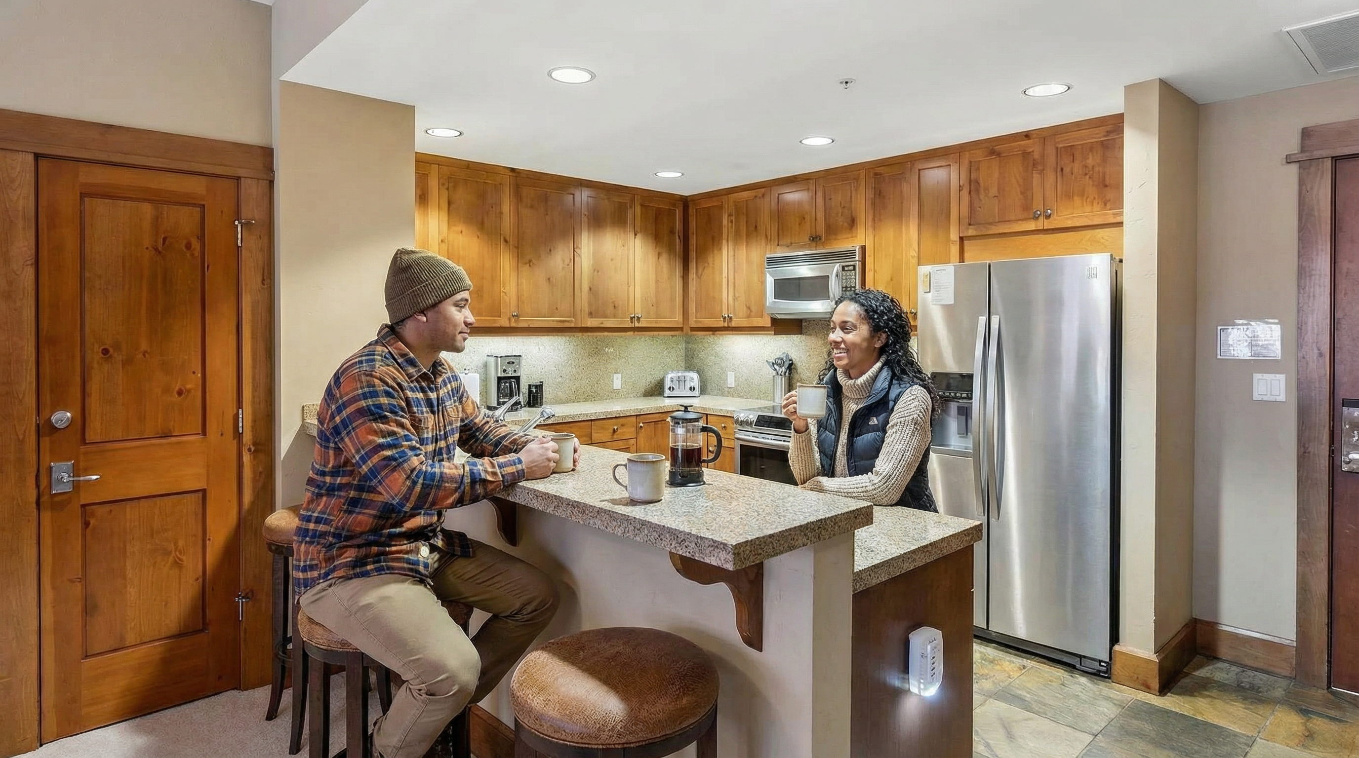 This is an interior shot of a kitchen. It features wooden cabinets, stainless steel appliances, and a granite countertop bar with seating. Two people are enjoying coffee at the bar, creating a warm and inviting atmosphere.