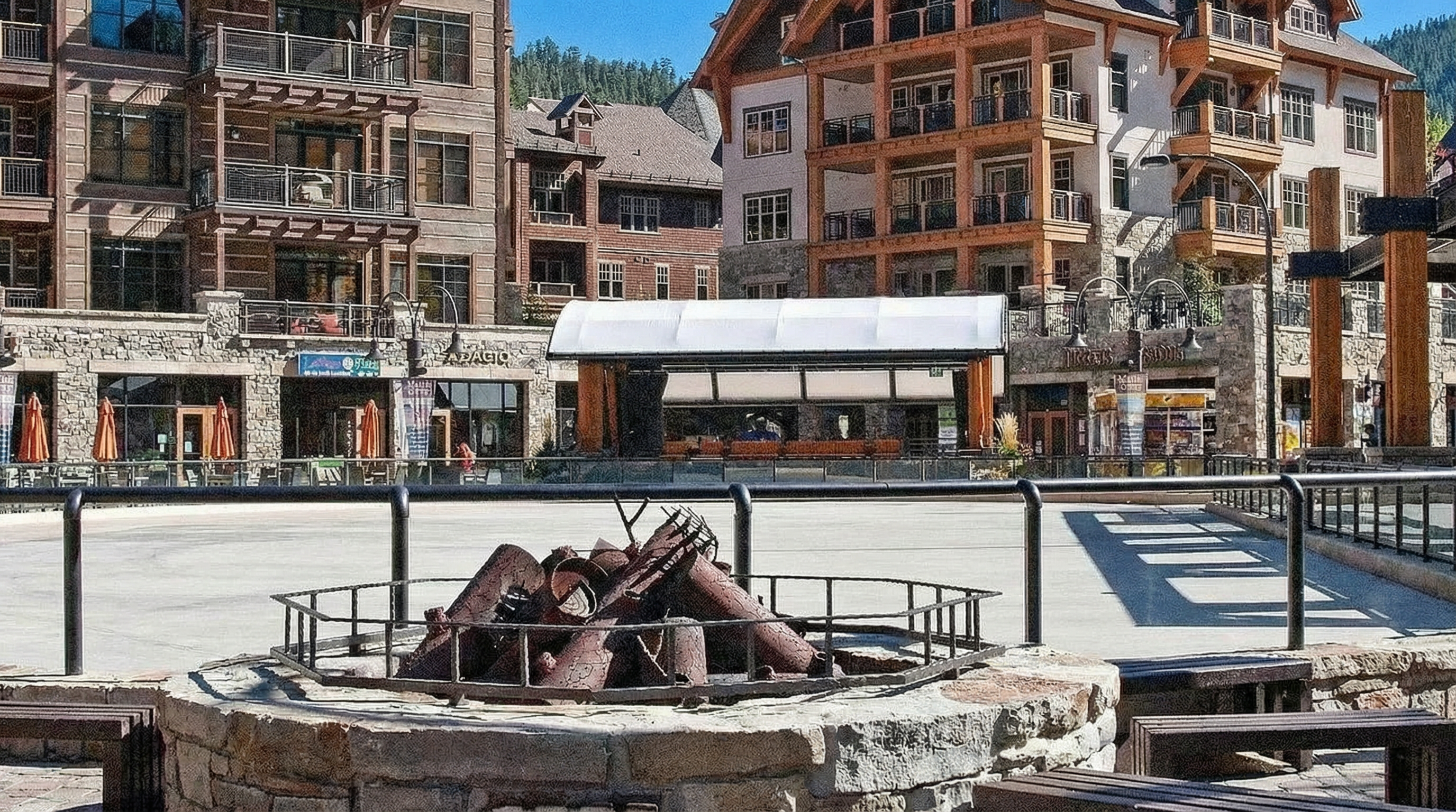This image showcases a community gathering space, likely a central plaza or common area. A prominent fire pit feature, surrounded by seating, anchors the space. The background features mixed-use buildings with retail spaces and residential units above, suggesting a vibrant, walkable neighborhood setting.