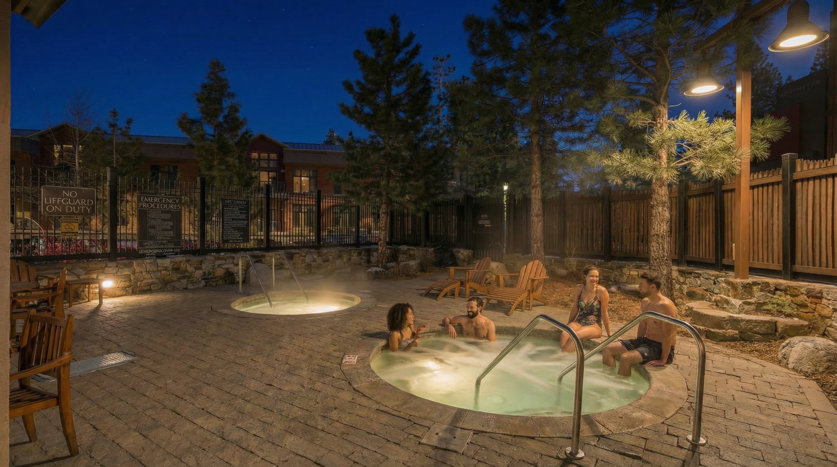 This image showcases an outdoor spa area at dusk. Several people are relaxing in the hot tub, and another smaller hot tub is visible nearby. The area is paved with stone and surrounded by trees and a wooden fence, creating a private and appealing amenity space.