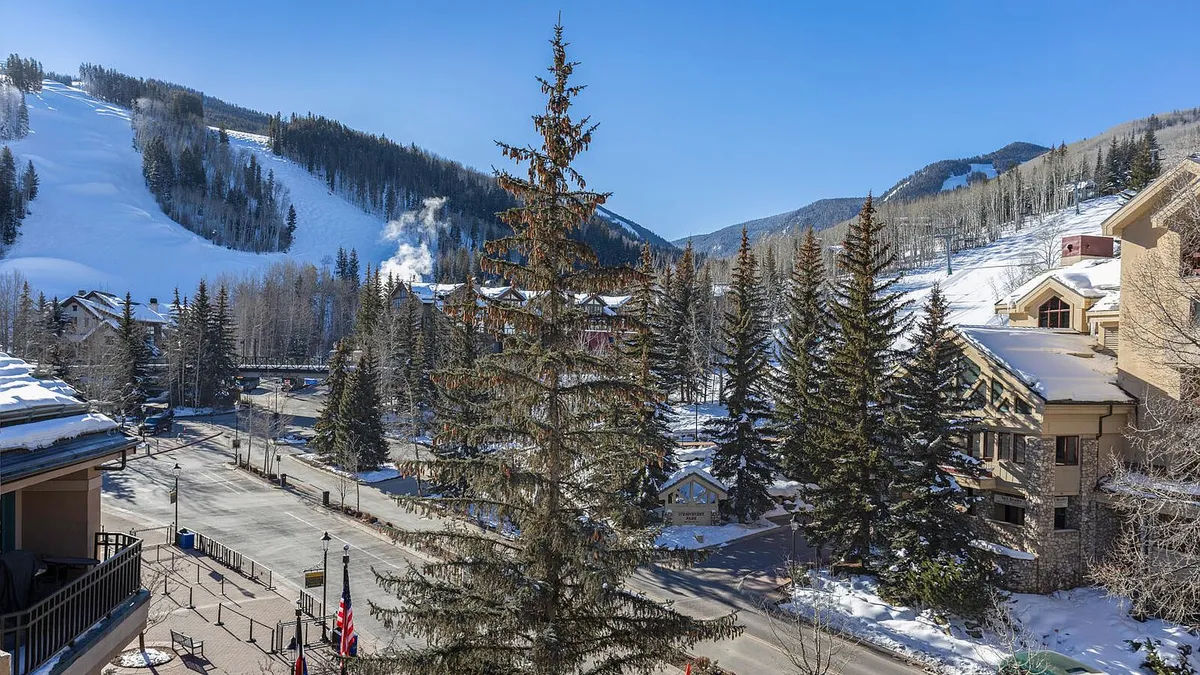 This image showcases a charming mountain resort area in winter. The street is lined with evergreen trees, flags, and quaint buildings, with snow-covered slopes visible in the background. The setting evokes a sense of luxury and recreation, ideal for a vacation property.