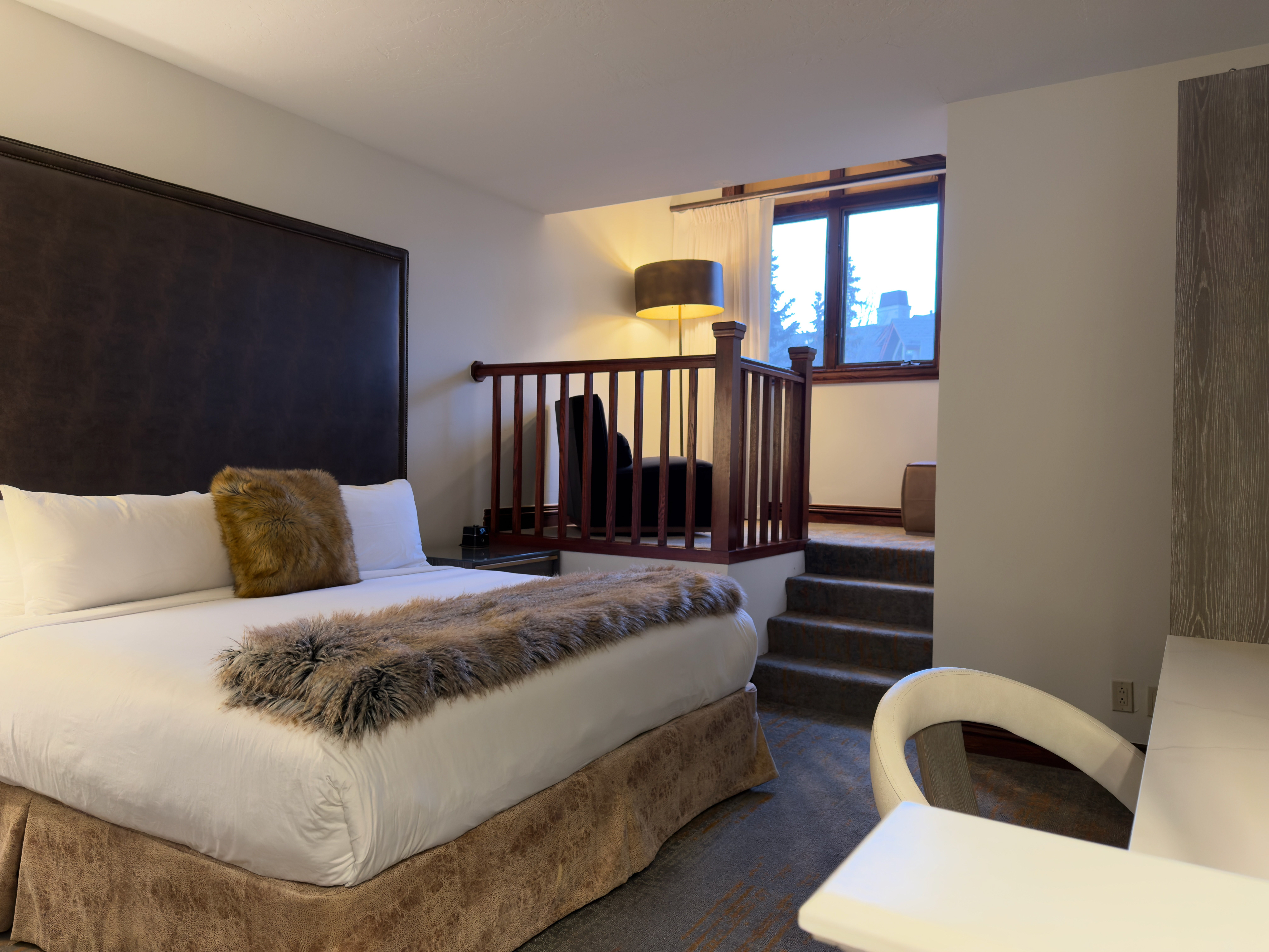 This bedroom has a dark leather headboard, a bed with white linens and a furry throw blanket. There is a loft area above the bed with a railing. The room is well-lit with a bedside table and an adjacent desk.