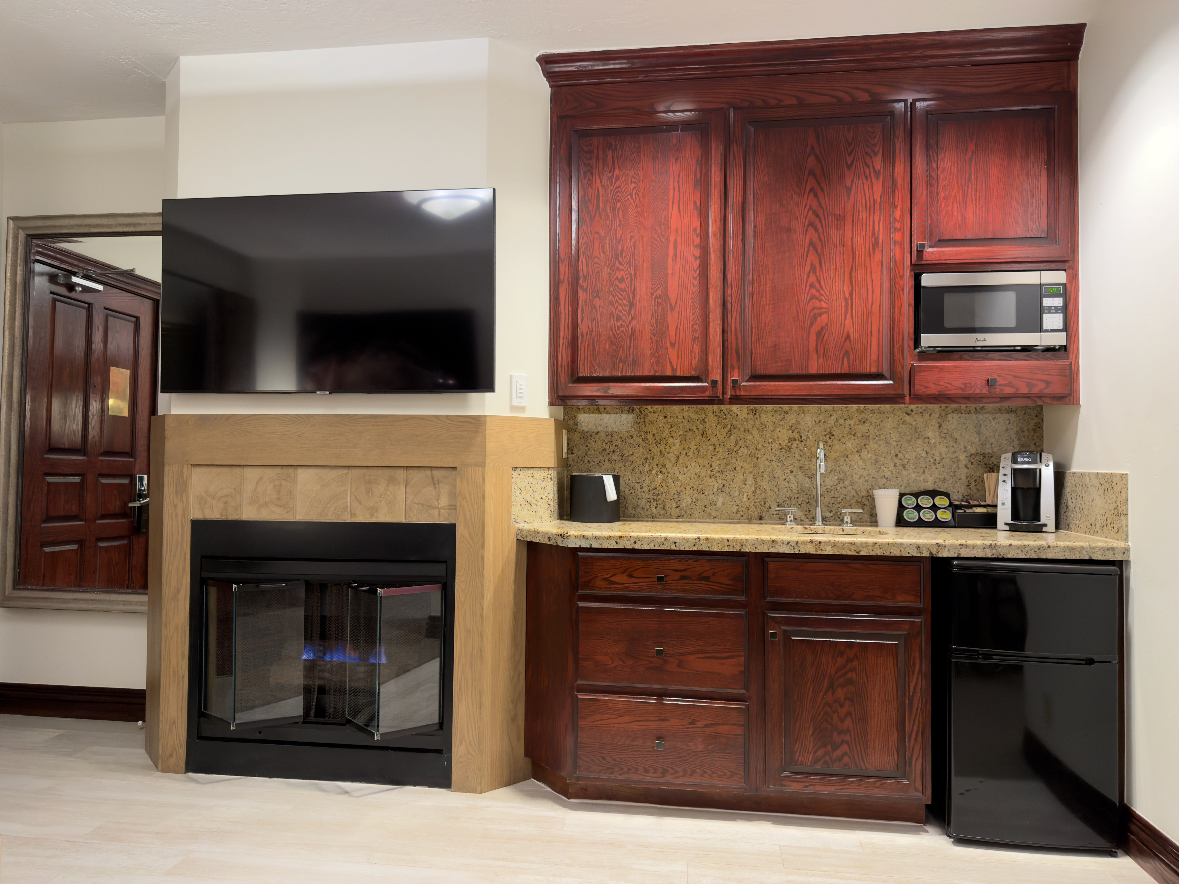 The image showcases a kitchenette area, possibly within a hotel suite or small apartment. Dark wood cabinetry, complemented by a tan countertop, provides storage space above and below. Appliances include a microwave, coffee maker, sink, and mini-fridge, indicating convenience and functionality for short-term or compact living.