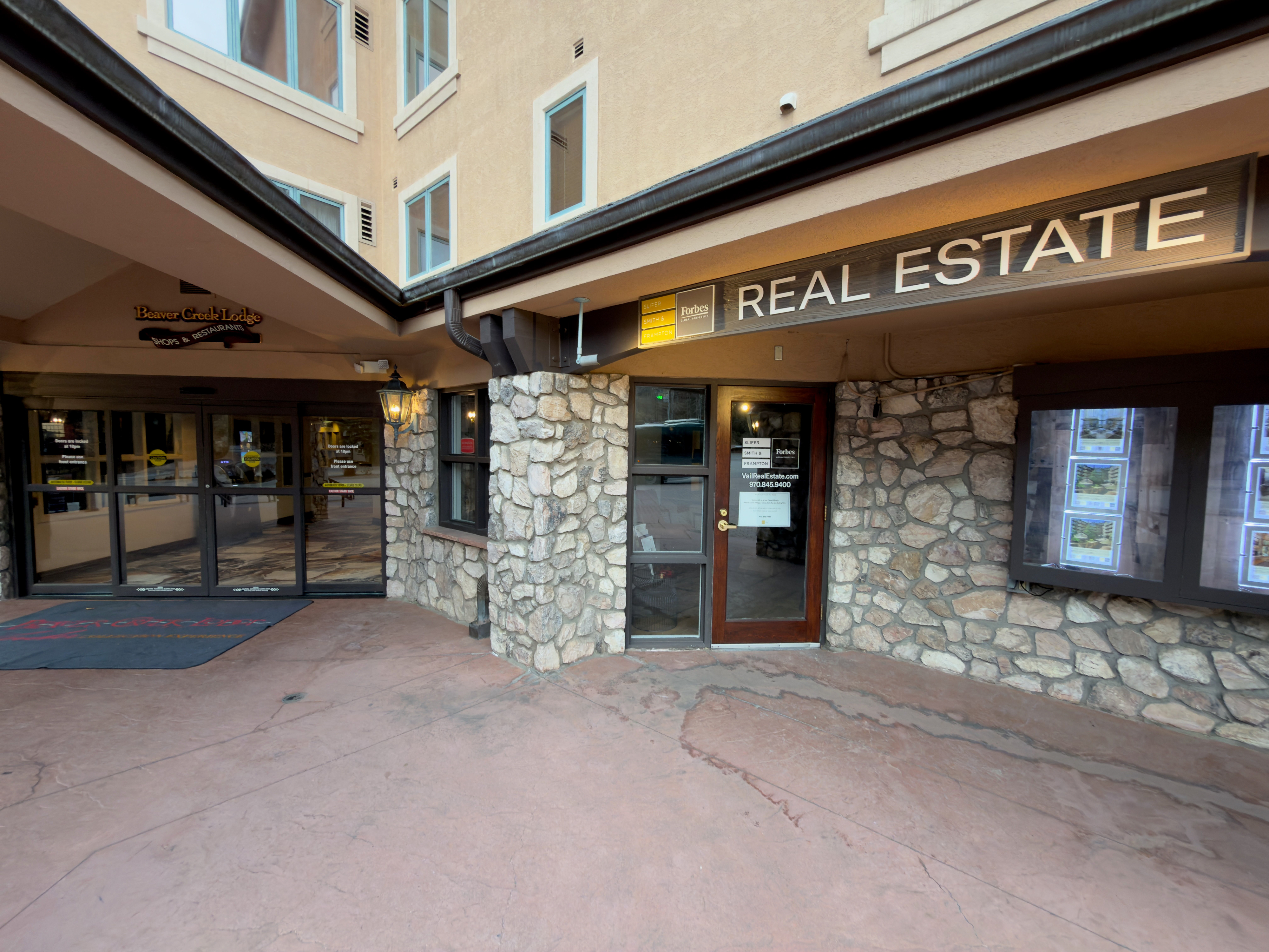The image shows the entrance to a real estate office located within Beaver Creek Lodge. The entryway features a stone facade, a 'REAL ESTATE' sign with Forbes logo above the door, and display windows showcasing property listings. The space is well-lit and appears inviting.