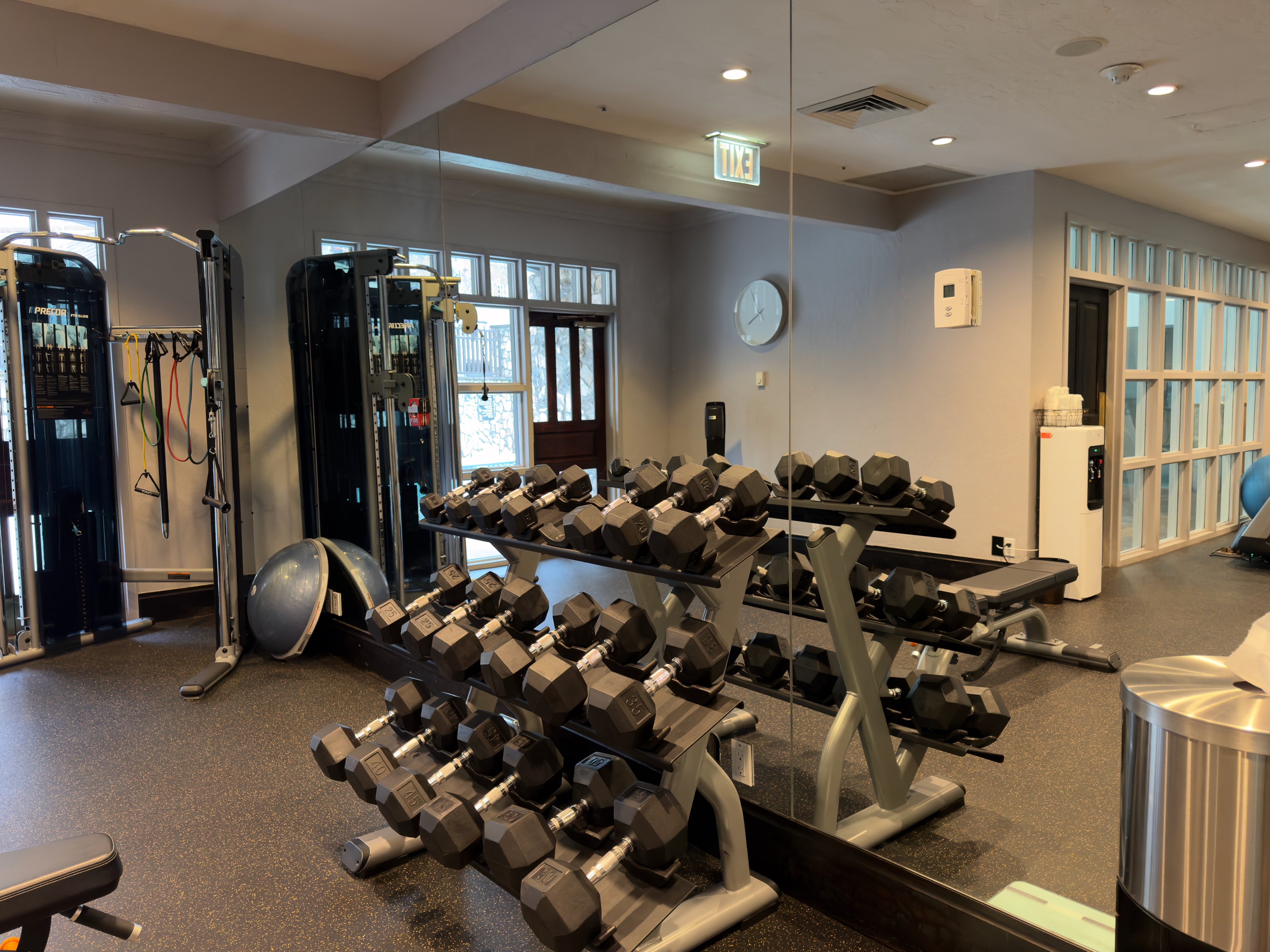 The image showcases a well-equipped home gym featuring weightlifting equipment, including racks of dumbbells of various weights. A multi-functional exercise machine is visible to the left, adding to the versatility of the space. The gym is situated indoors with a mirrored wall that enhances the sense of space and allows for form observation during workouts.