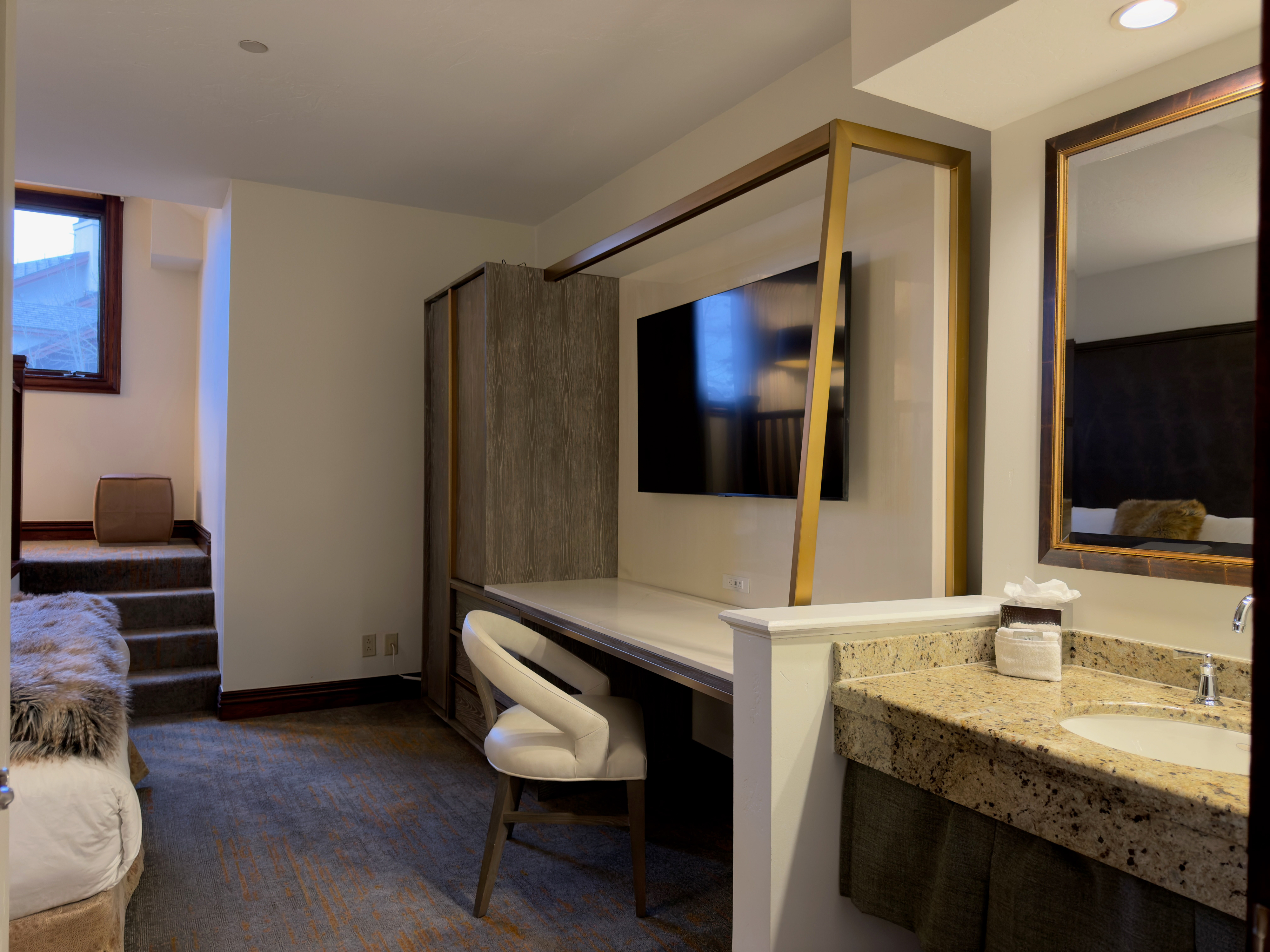 The image showcases a well-appointed guest bedroom featuring a desk area with a mounted television and a chair. A mirrored bathroom vanity area sits adjacent, and stairs lead up to what appears to be another sleeping area. The room utilizes neutrals and muted tones to create a calming and inviting atmosphere, suitable for a comfortable stay.