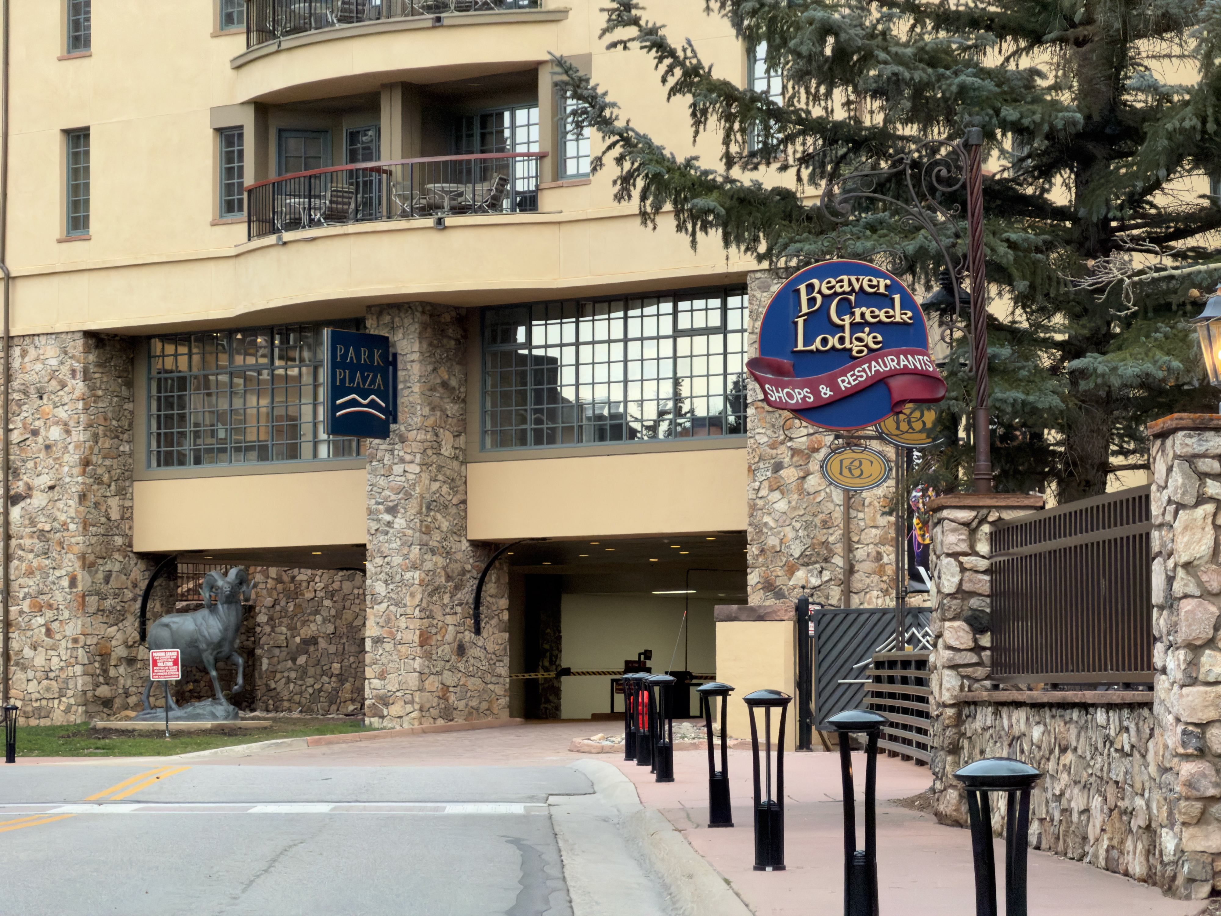 This is a front exterior view of the Beaver Creek Lodge. It features a stone facade on the lower levels, a beige upper level, and prominent signage for the lodge's shops and restaurants. There is an entrance to what appears to be a parking garage under the building.