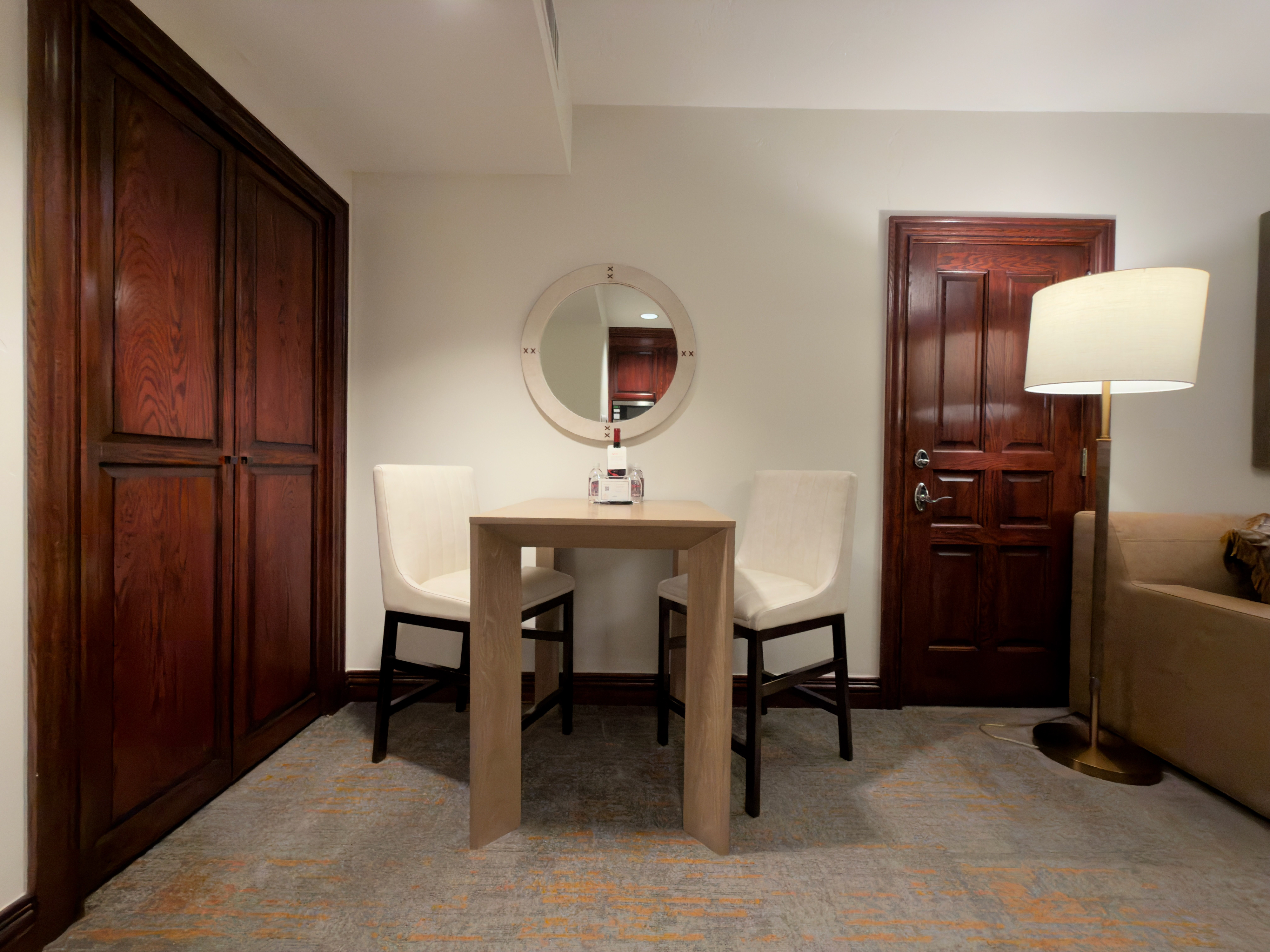 This interior shot showcases a small dining area, possibly within a hotel room or small apartment. A square table with two chairs sits in the center of the room, with a round mirror on the wall above. Dark wood accents on the door and closet add a touch of traditional elegance.