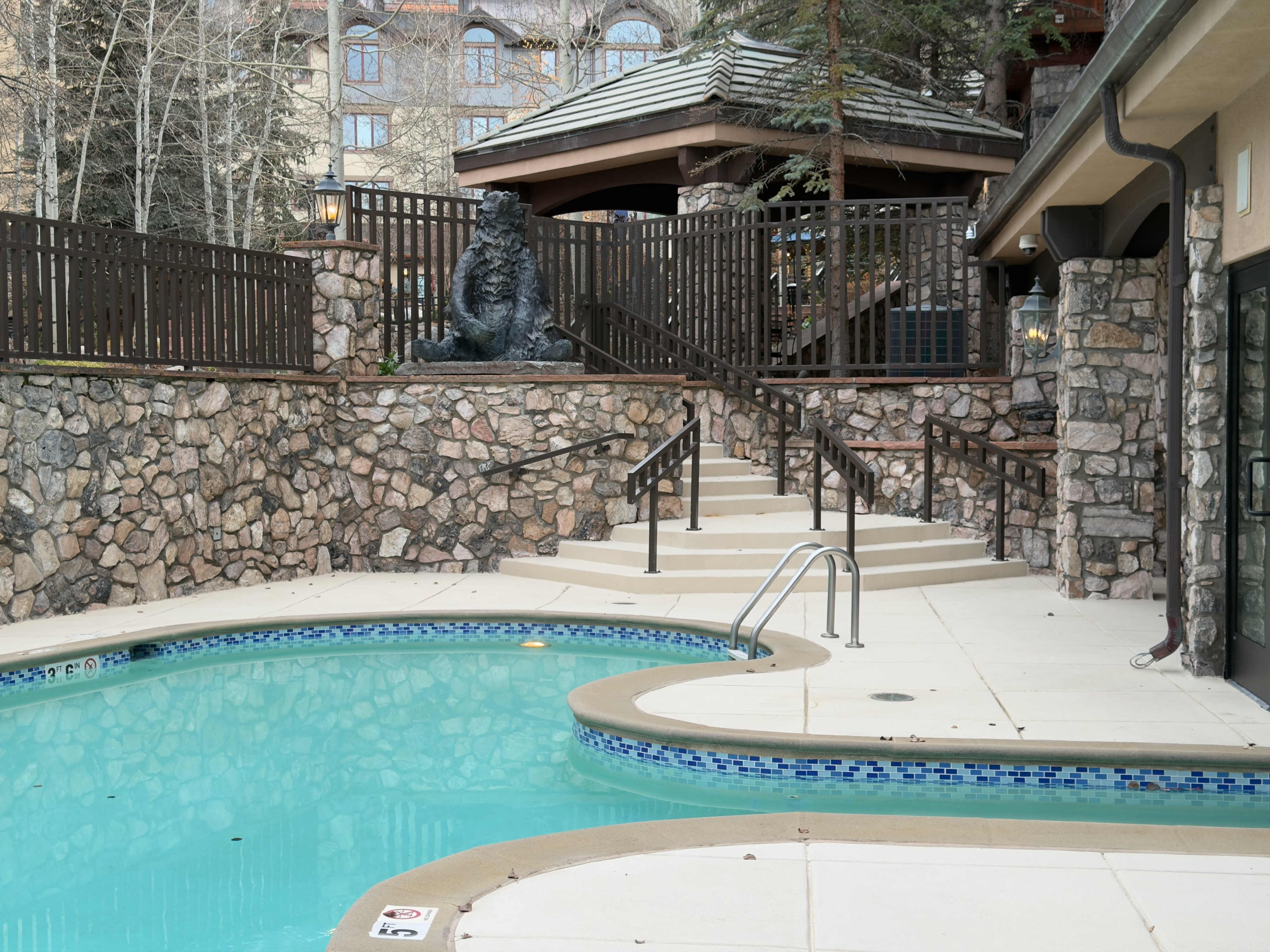 This image showcases a well-maintained outdoor pool area, likely belonging to a luxury property. The pool is turquoise and surrounded by a concrete patio. Stone walls, tasteful landscaping, and a decorative bear statue add to the upscale ambiance, potentially enhancing the property's appeal for relaxation and entertainment.