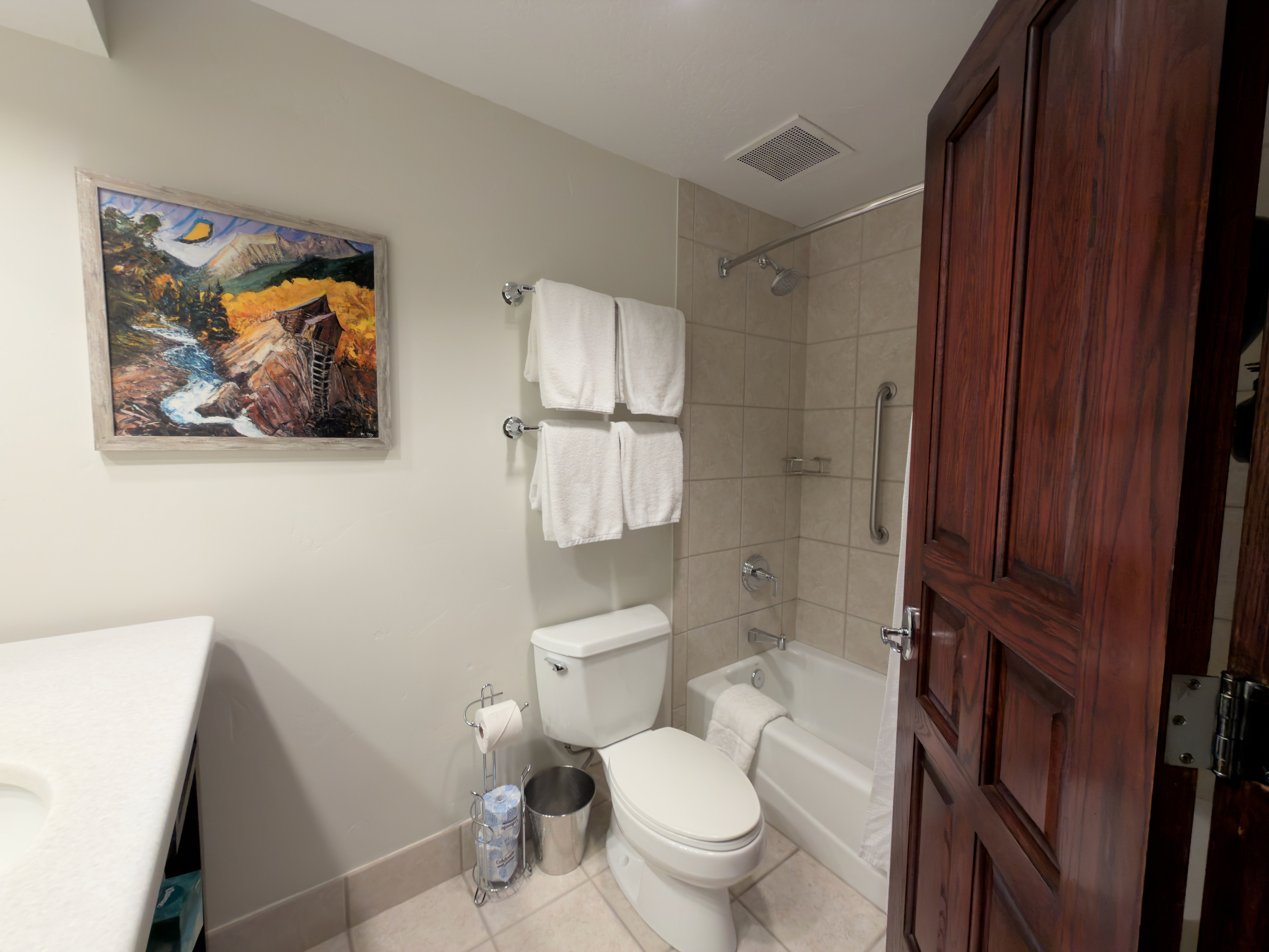 This is a bathroom featuring neutral-toned tiles in the shower/tub area, a standard white toilet, and towel racks with white towels. A framed painting hangs above the toilet, and a dark wood door leads into the bathroom. The space appears functional and well-maintained.
