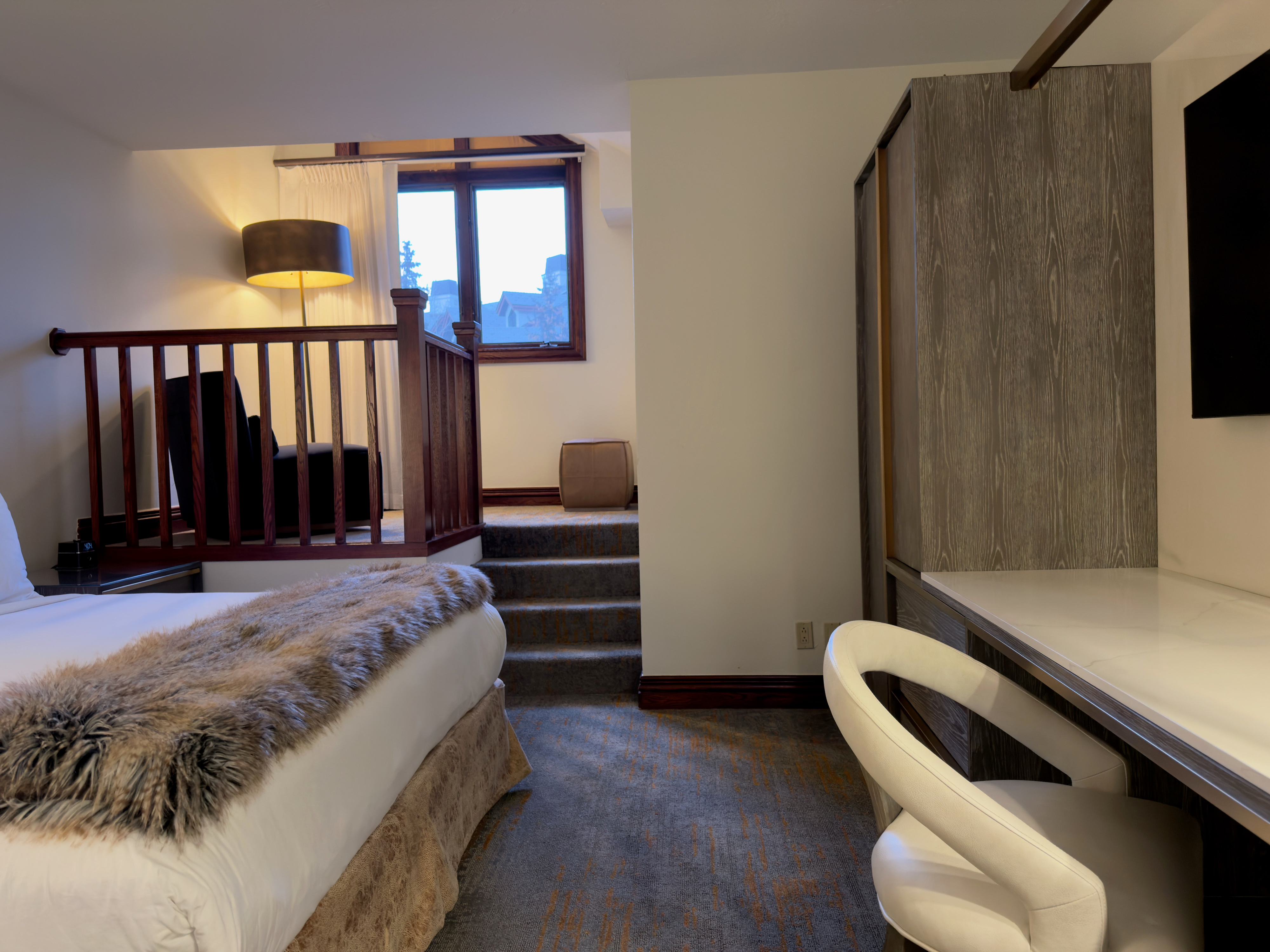 This is an interior shot of a bedroom. It features a bed with a decorative fur throw, a small staircase leading to a seating area with an armchair, and a desk area with a white chair. The room has a modern, cozy feel, with a focus on comfort and style.