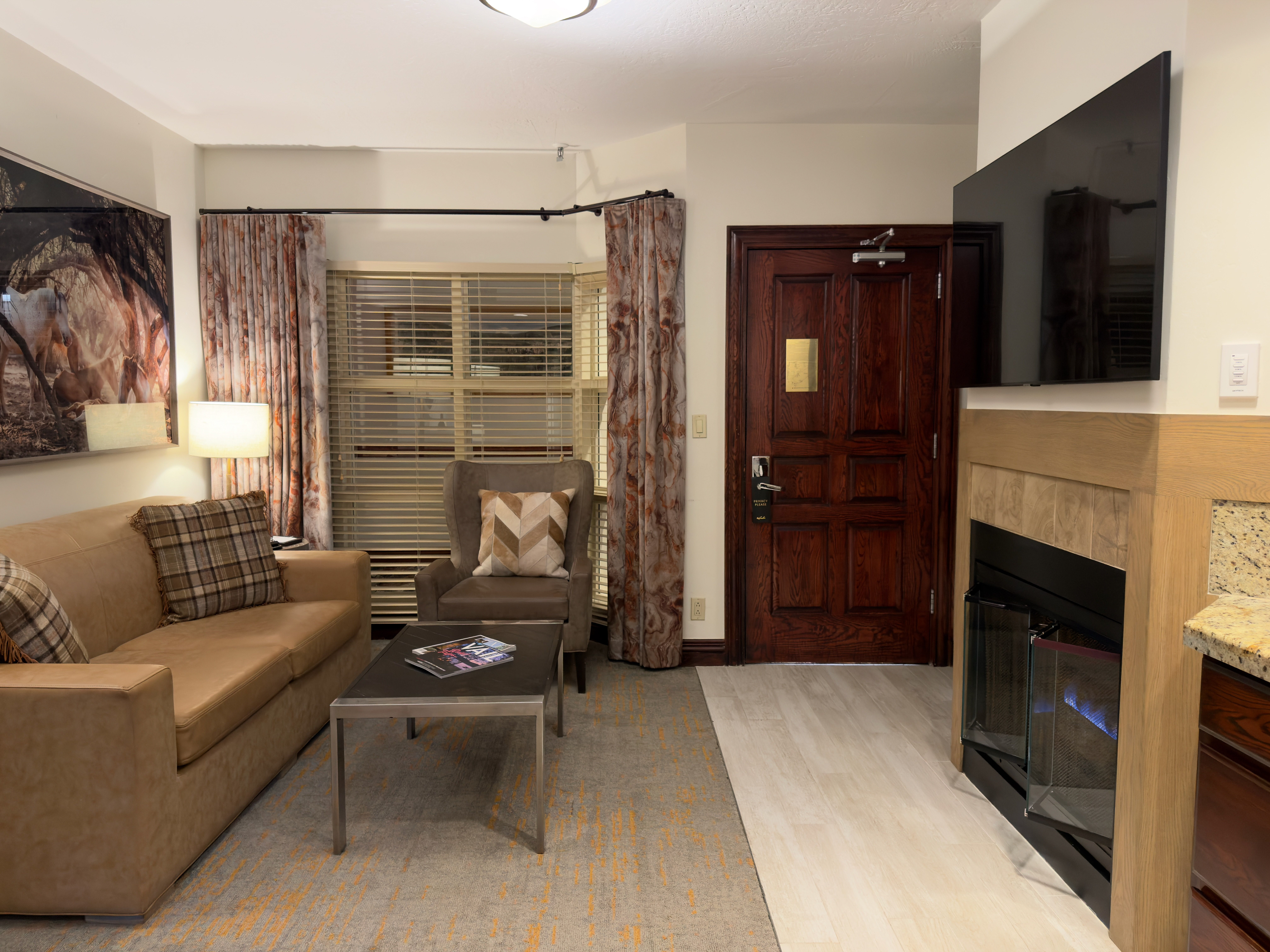 This interior shot showcases a cozy living room featuring a tan leather sofa adorned with plaid cushions, an armchair, and a modern coffee table. The room is decorated with curtains flanking a window and an artwork above the sofa. A fireplace mantel with a mounted television adds a touch of contemporary living, suggesting a comfortable and inviting space for relaxation and entertainment.