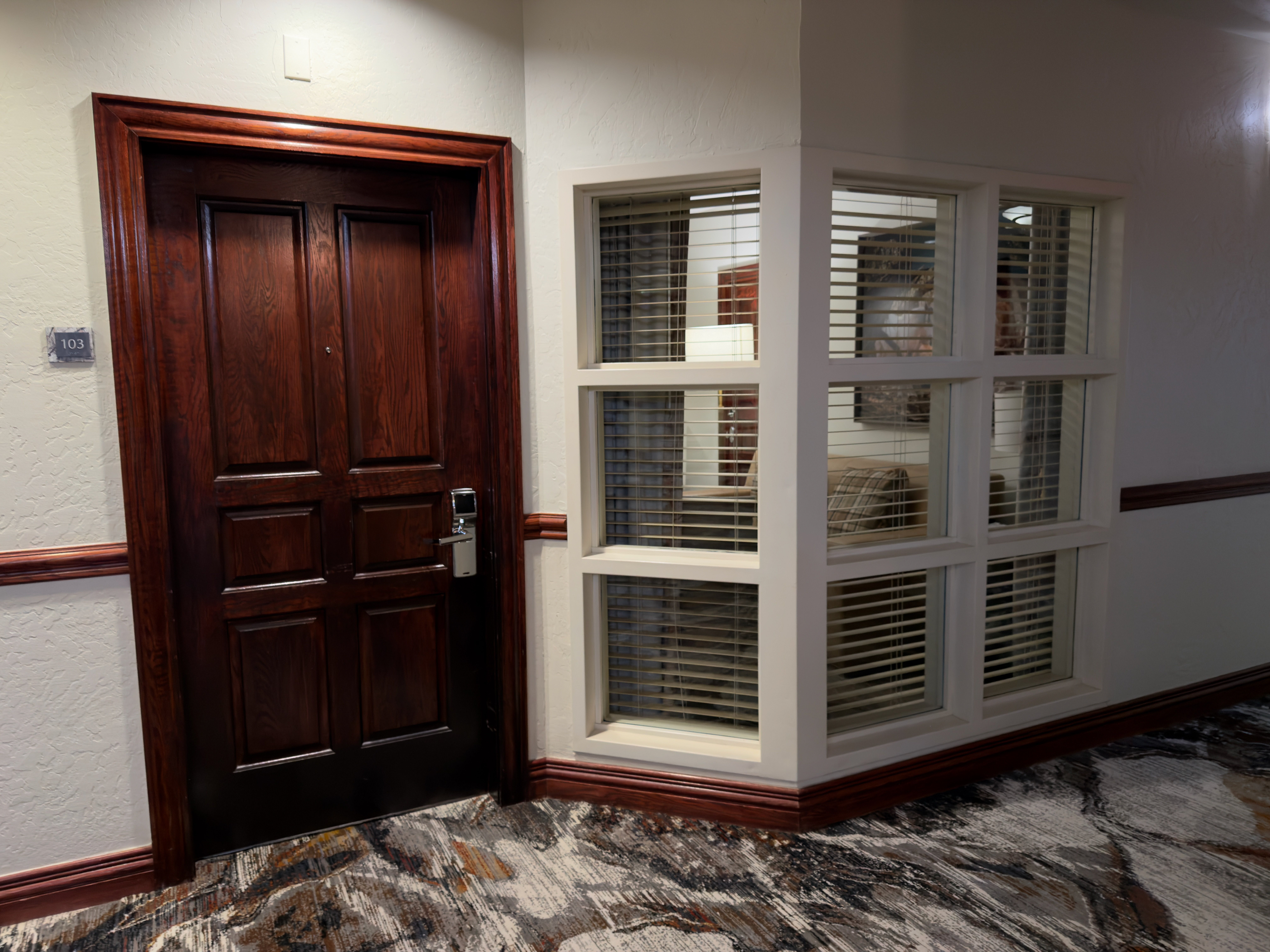 This is a hallway featuring a dark wood door with room number signage. Next to the door is a bay window with closed blinds, suggesting privacy and controlled light. The hallway has patterned carpeting adding a modern touch.