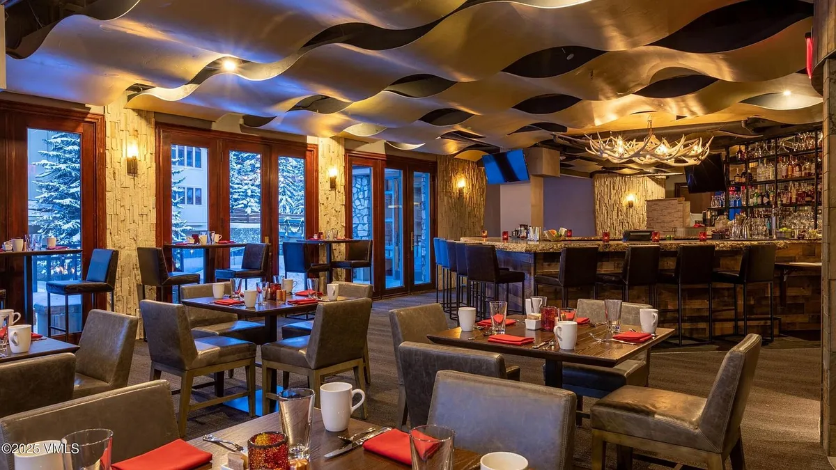 The image showcases a well-appointed dining area with multiple set tables and chairs. The room features unique ceiling decor, stone accent walls, and large windows providing natural light. A bar area is visible in the background, complete with a full liquor selection and antler chandelier, enhancing the upscale ambiance.