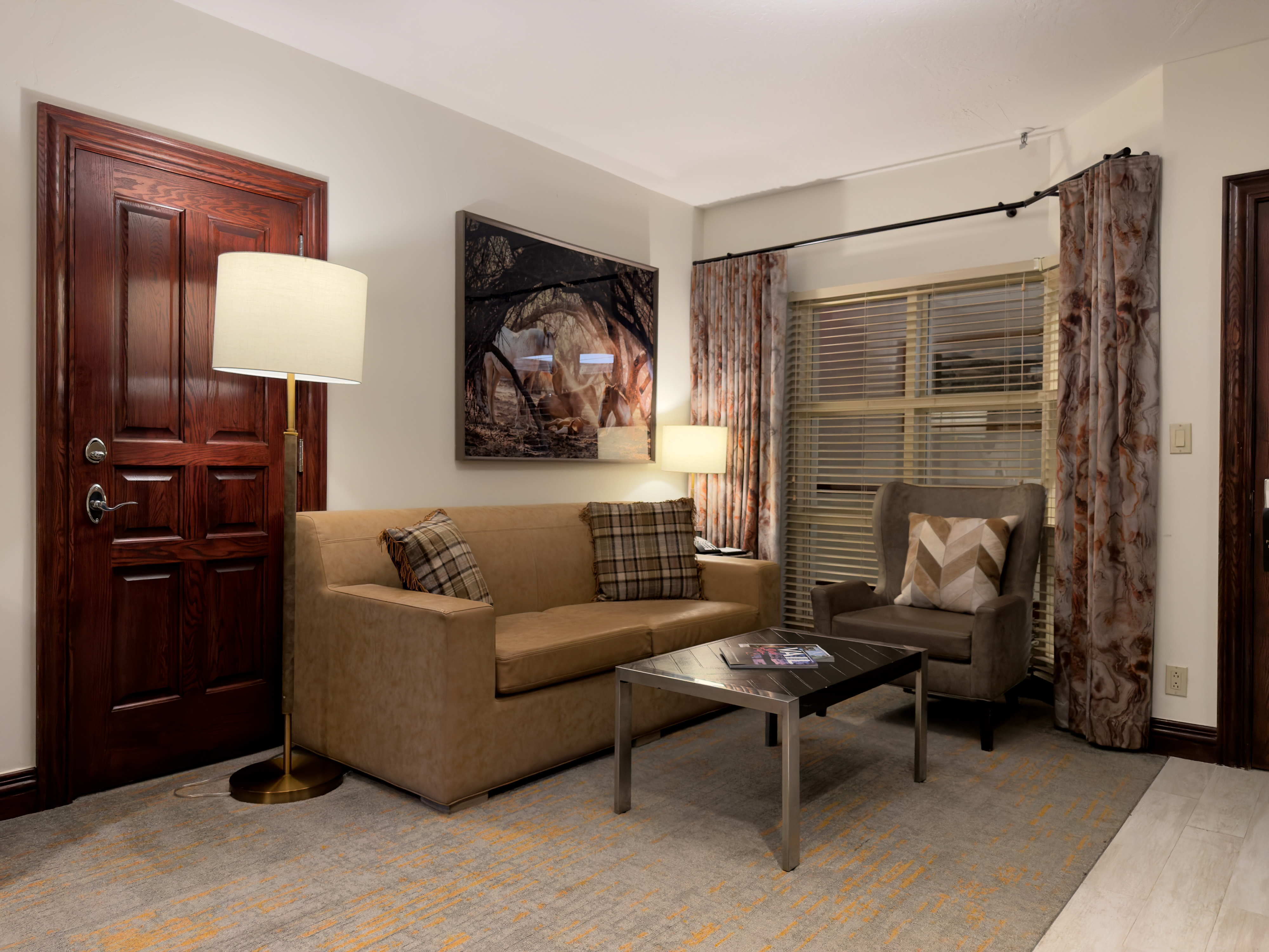 The image showcases a well-decorated living room. It features a tan sofa with plaid decorative pillows, an armchair with a patterned throw pillow, and a dark-topped coffee table with silver legs. The room is illuminated by a floor lamp and a table lamp, creating a cozy and inviting atmosphere.