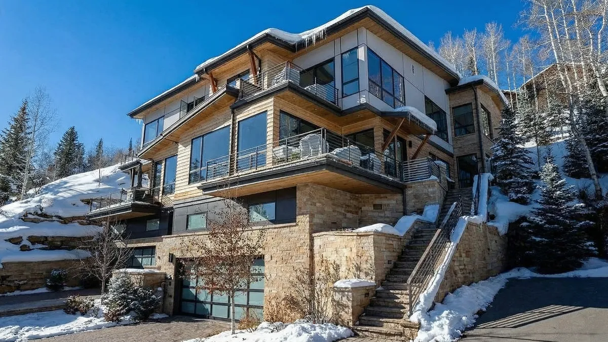 This is a front view of a modern, multi-story house with a stone base and wood and white siding. The house features large windows and balconies with metal railings. The presence of snow suggests a mountain or ski resort location, adding to the property's appeal.