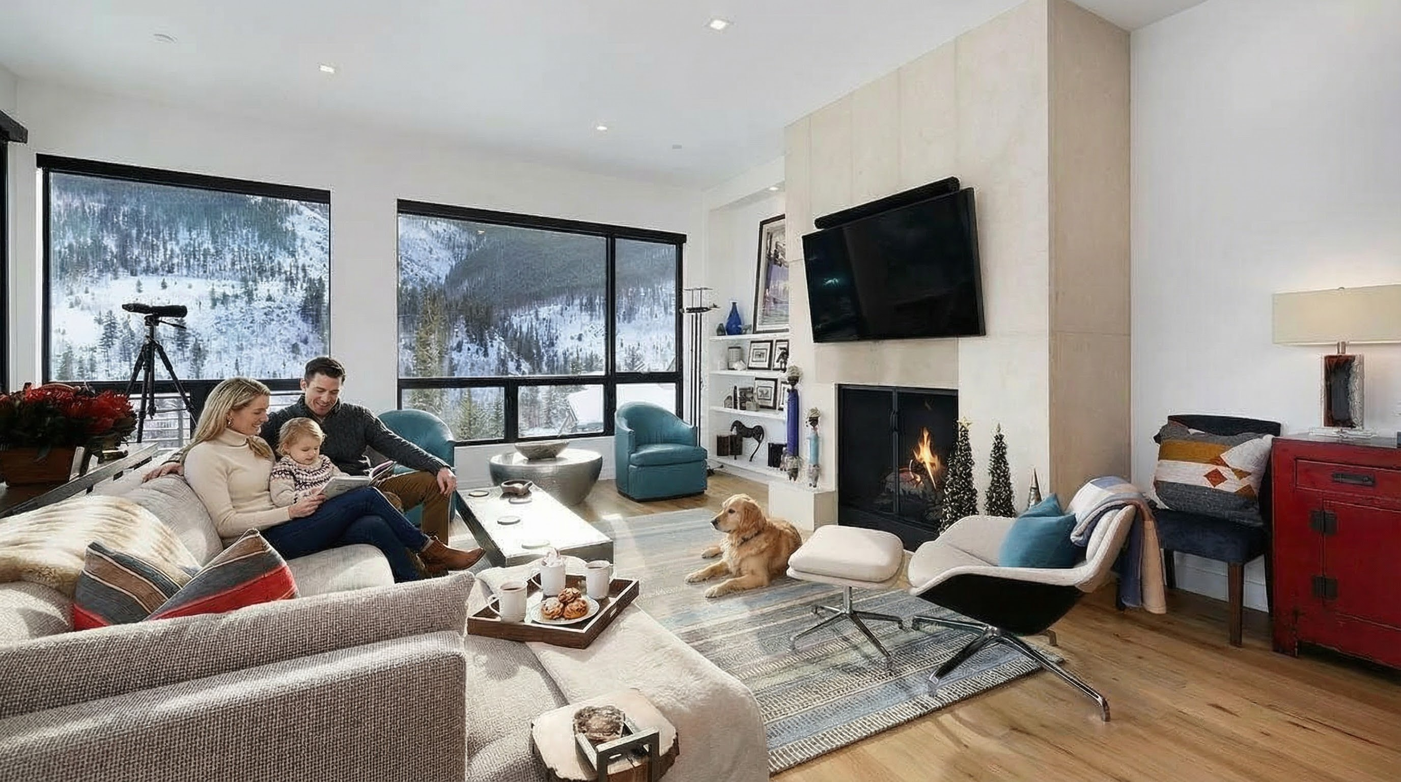 This is a well-lit living room featuring large windows with views of a snowy mountain landscape. The room's focal point is a modern fireplace with a mounted flat-screen television above it. A comfortable sectional sofa, a neutral rug, and stylish accent chairs create an inviting and cozy atmosphere.