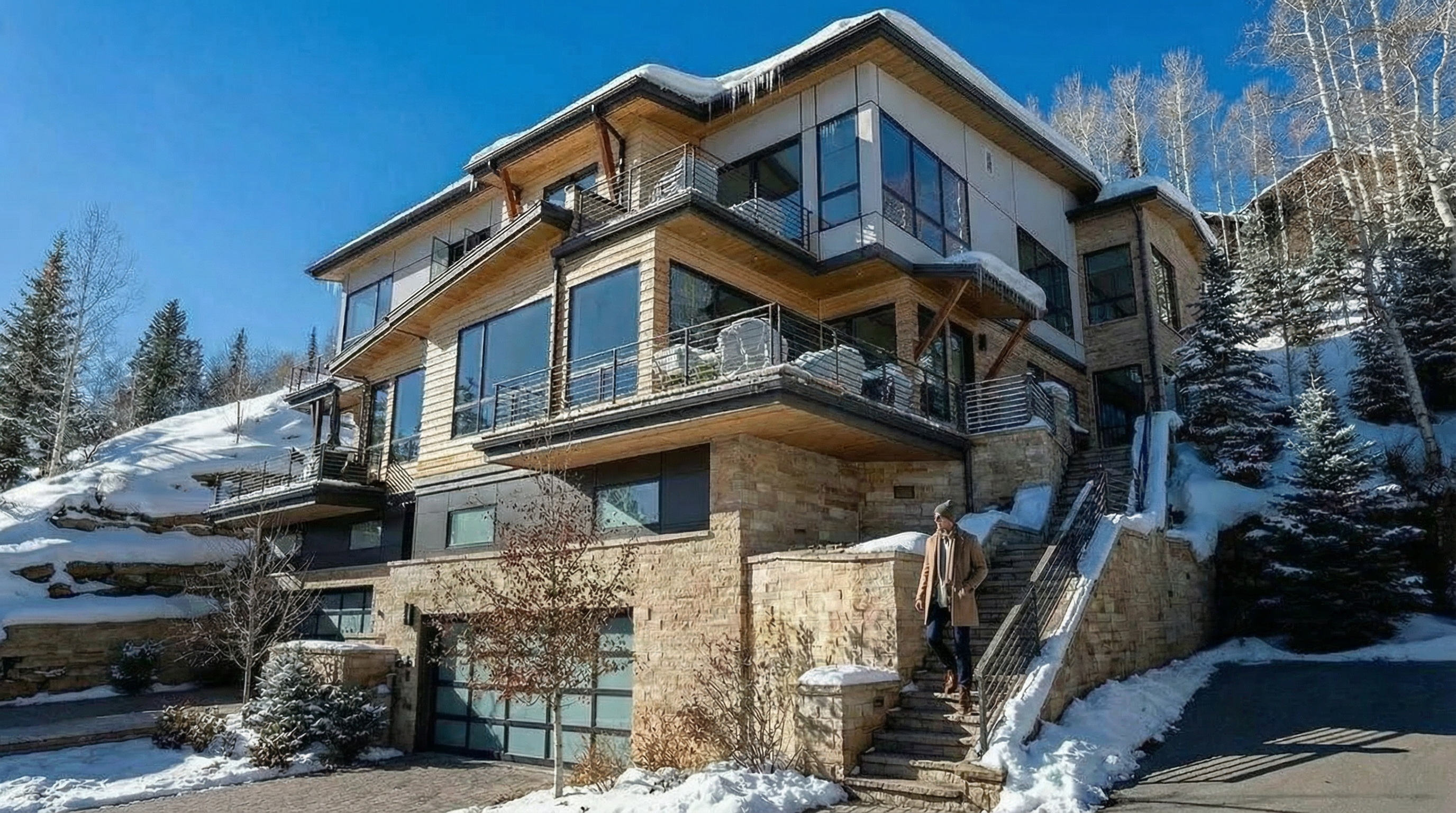 This is a stunning exterior shot of a multi-level home, likely located in a snowy, mountainous region. The design blends modern elements with natural materials like wood and stone, featuring large windows and balconies that offer impressive views. A stairway leads up to the residence, giving a sense of grandeur and sophistication.