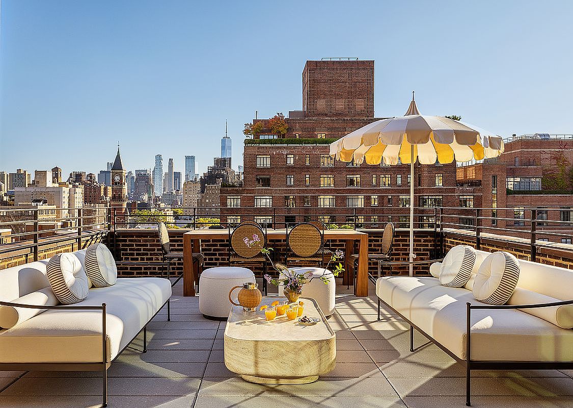 This image showcases a stylish rooftop patio or deck with elegant outdoor furniture. The space includes comfortable seating areas with plush cushions, a dining table with wicker chairs, and a chic coffee table with drinks and floral arrangements. An umbrella provides shade, while the cityscape offers a stunning backdrop, creating an inviting and luxurious outdoor living space.