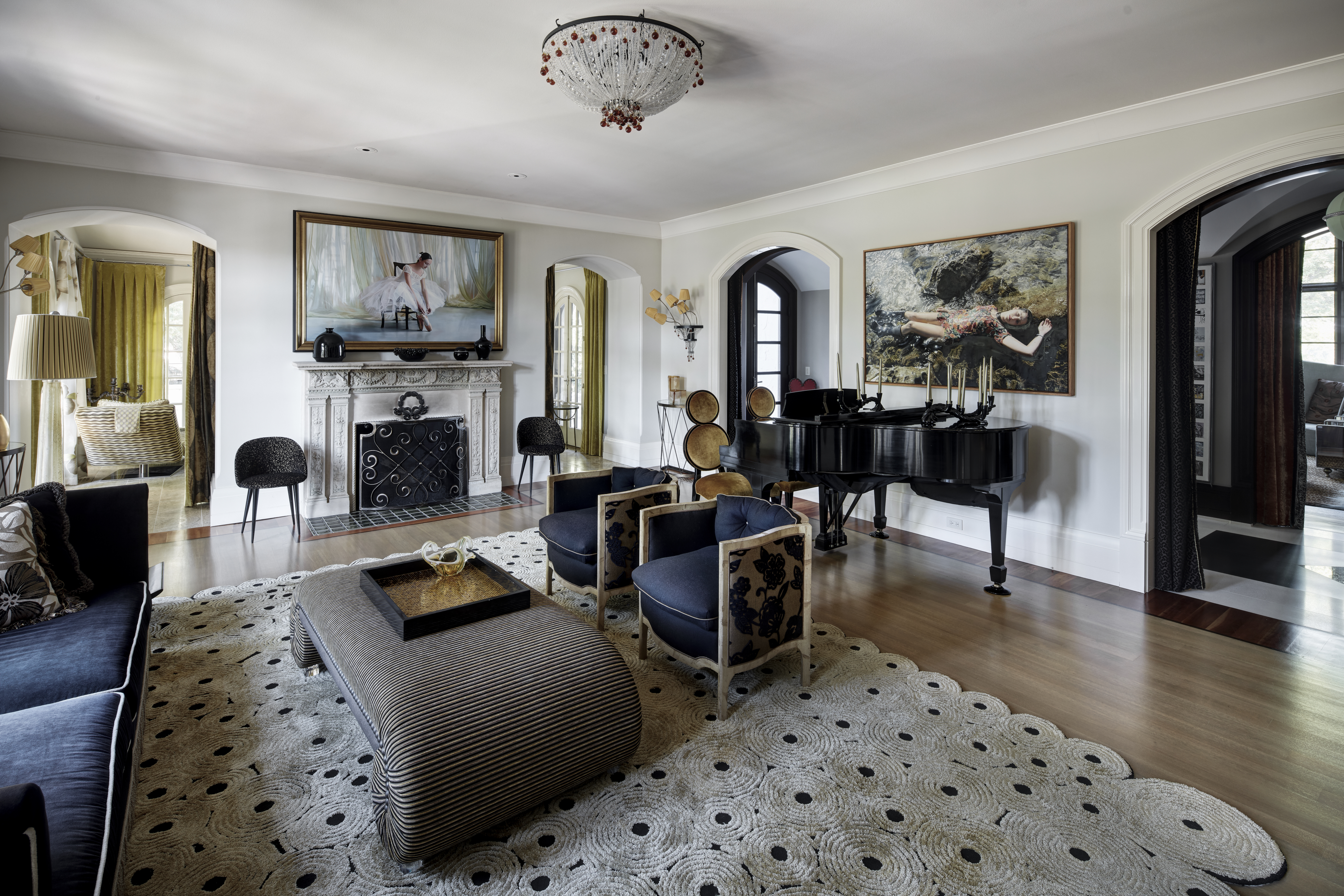This elegant living room features a grand piano, a fireplace with a large painting above it, and comfortable seating arranged on a patterned rug. The room is well-lit by a chandelier and natural light, creating a sophisticated and inviting atmosphere. Arched doorways lead to other areas of the home, enhancing the sense of spaciousness and flow.