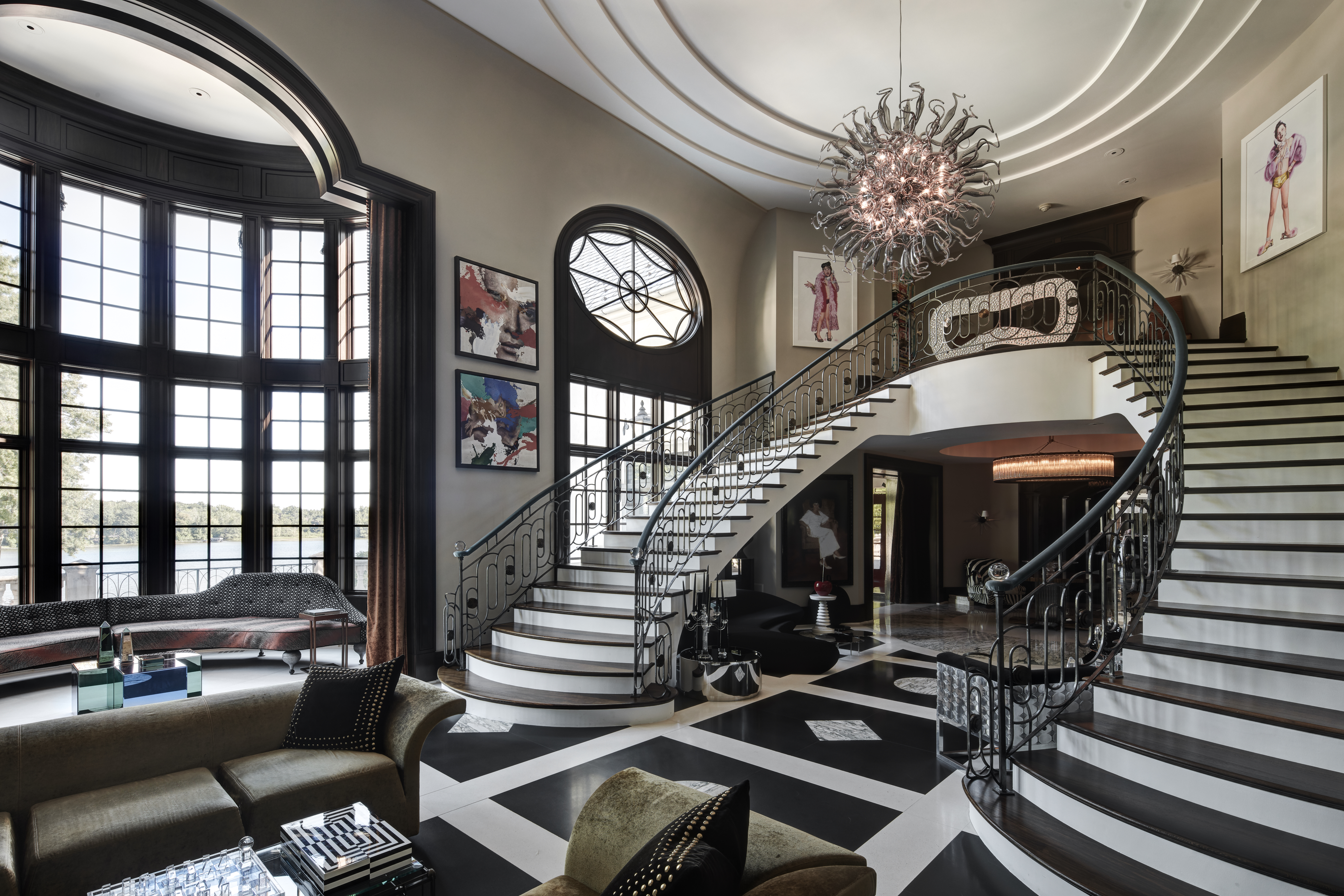 This grand foyer features a sweeping staircase with ornate iron railings and contrasting dark wood and white steps. The black and white patterned marble flooring adds a touch of elegance, while a large, modern chandelier illuminates the space. The room is filled with natural light from the large windows, creating a bright and inviting atmosphere.