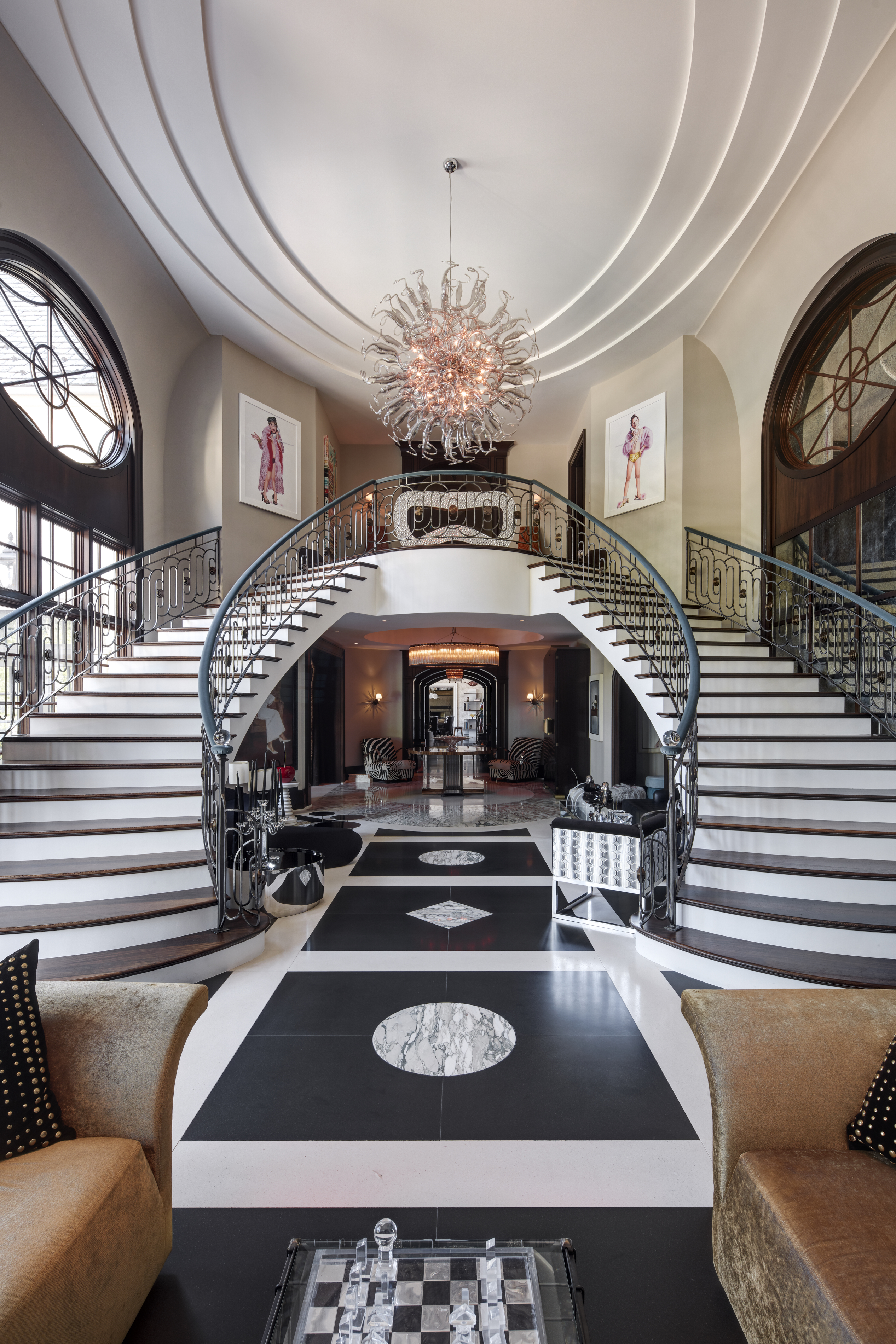 This grand foyer features a dramatic dual staircase with ornate iron railings, leading to an upper level. A striking chandelier hangs from the ceiling, illuminating the black and white patterned floor. Artwork adorns the walls, and the space is furnished with elegant seating, creating a luxurious and inviting entrance.