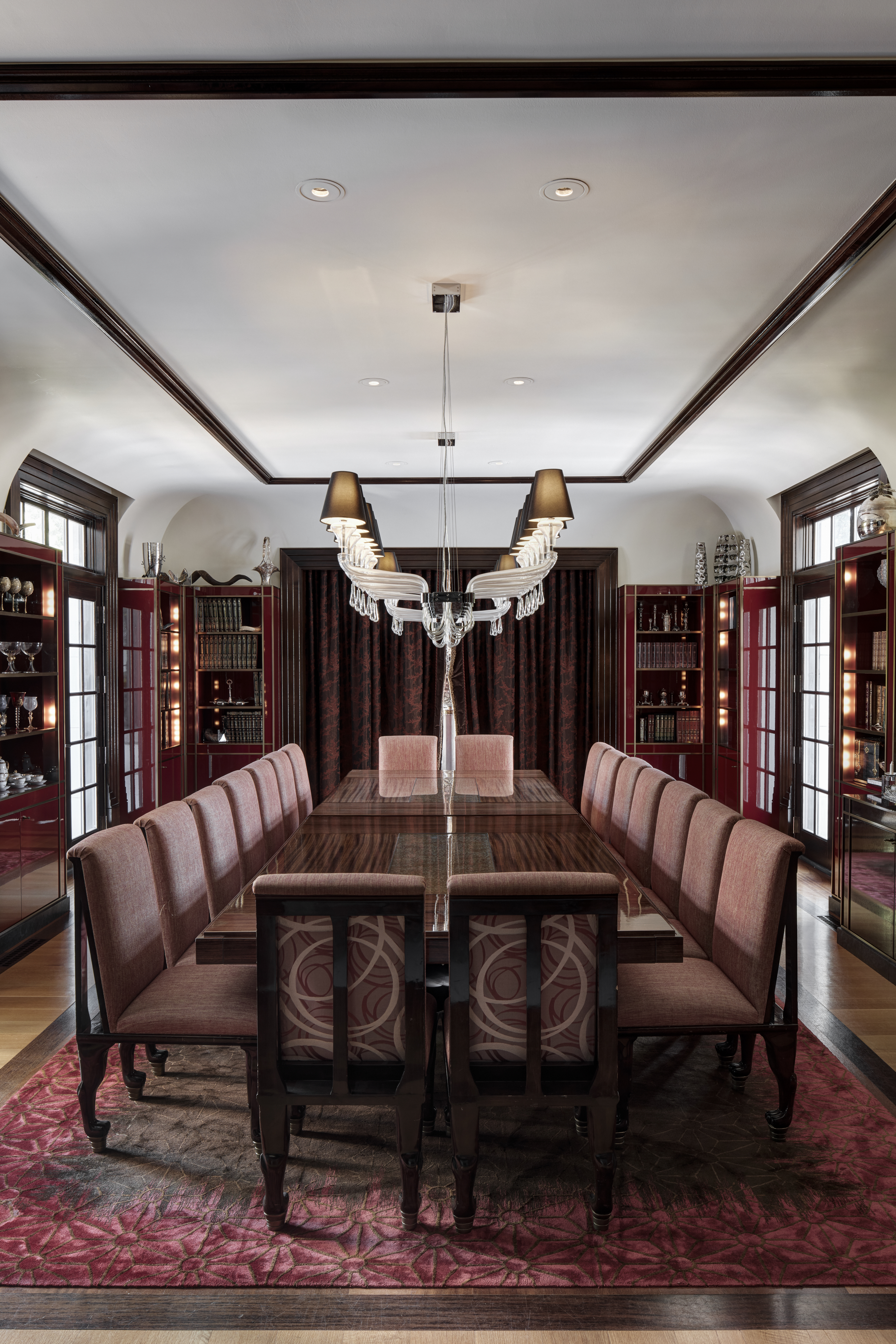 This is a formal dining room featuring a long, polished wood table with seating for many. The room is decorated with a large chandelier, built-in cabinets displaying glassware, and a patterned rug. The overall impression is one of elegance and sophistication, suitable for large gatherings and formal dining.