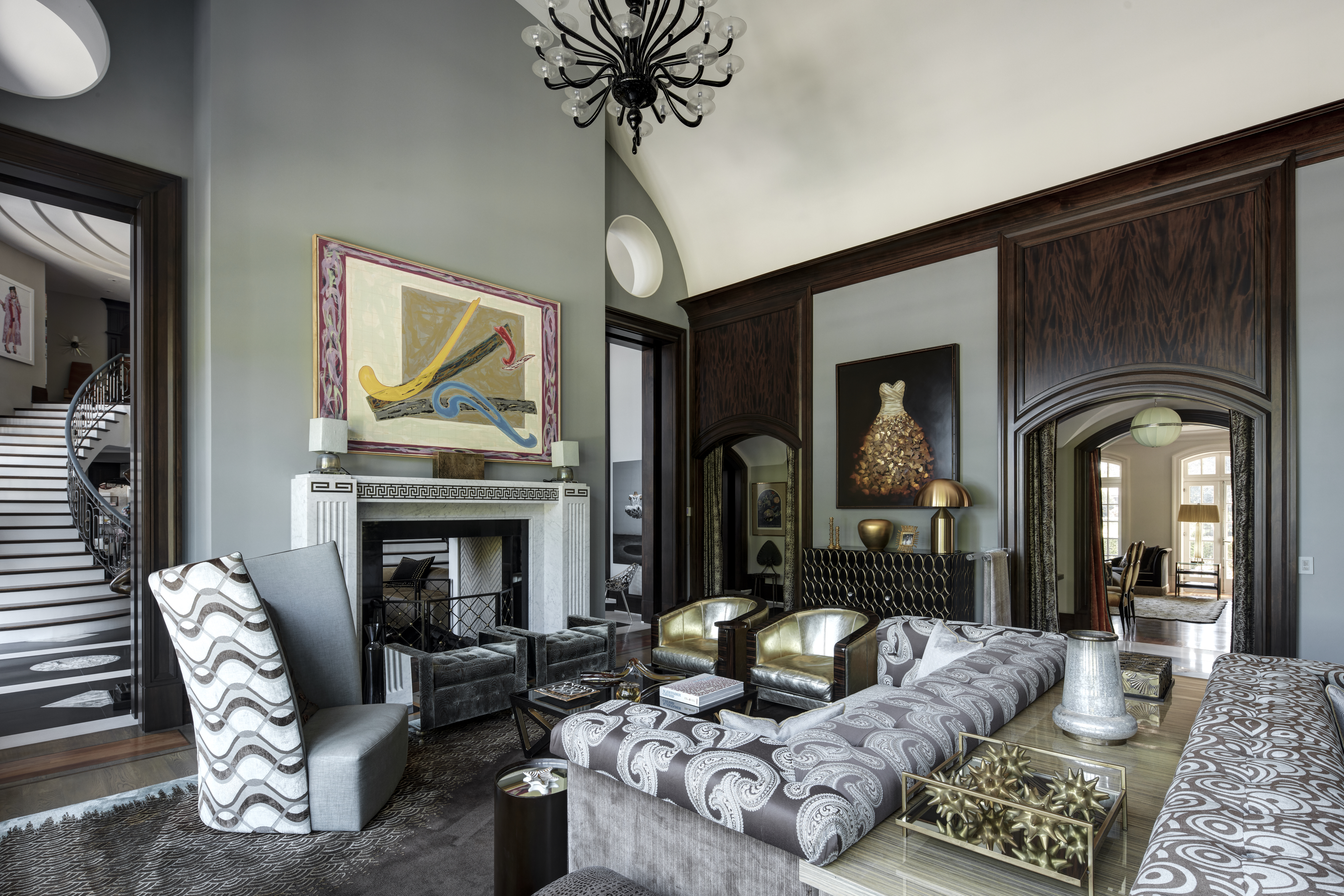 This is an interior shot of a luxurious living room featuring a sophisticated design with a blend of modern and classic elements. The room includes a fireplace with a marble surround, unique artwork, and stylish seating arrangements with patterned sofas and armchairs. Dark wood paneling adds a touch of elegance, while the overall ambiance is one of refined comfort and upscale living.