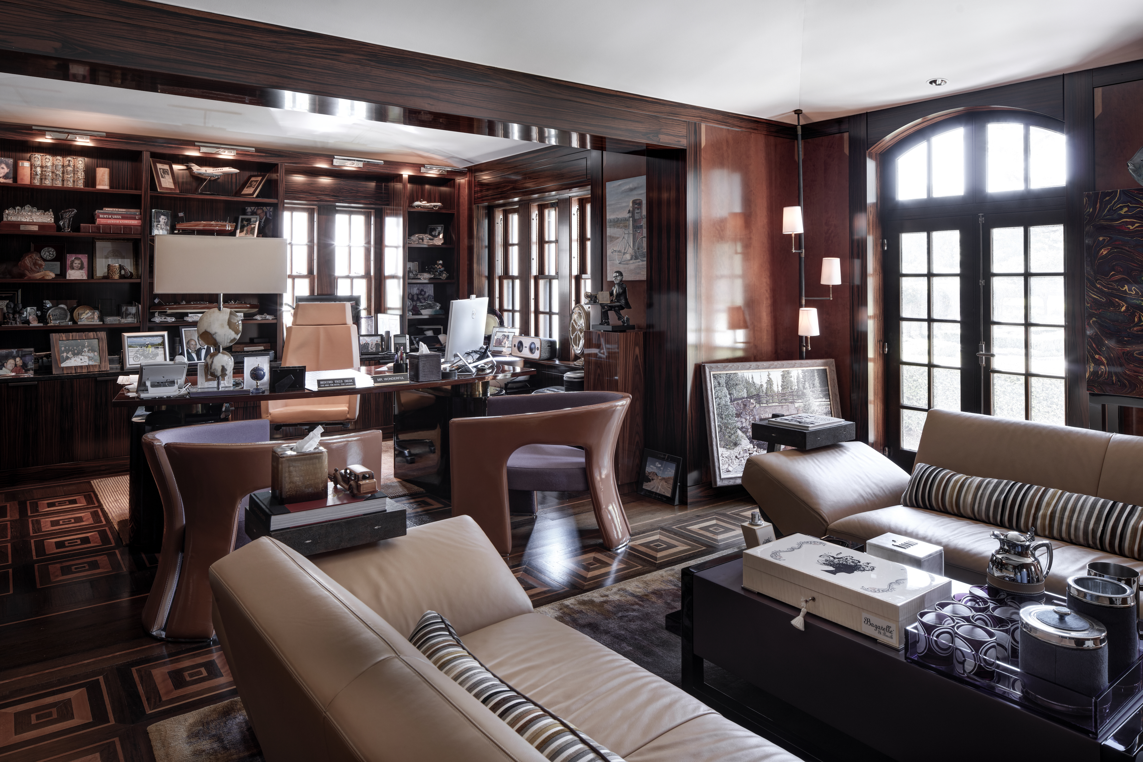 This luxurious office space features rich, dark wood paneling and built-in bookshelves, creating a sophisticated and professional atmosphere. The room is furnished with a large executive desk, comfortable leather seating, and stylish decor, including artwork and decorative objects. Natural light streams in through the windows, enhancing the warm and inviting ambiance.