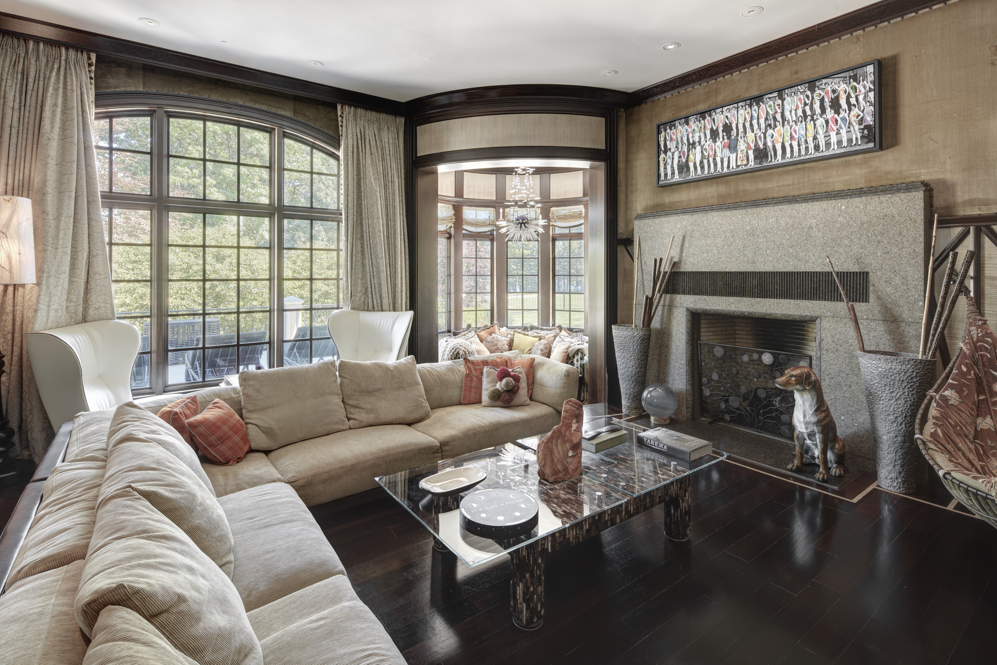 This is a luxurious living room featuring a large sectional sofa, a glass-topped coffee table, and a stone fireplace with decorative elements. The room is well-lit by a large arched window and a bay window, offering views of the outdoors. The dark wood flooring and rich wall textures contribute to an elegant and sophisticated atmosphere.