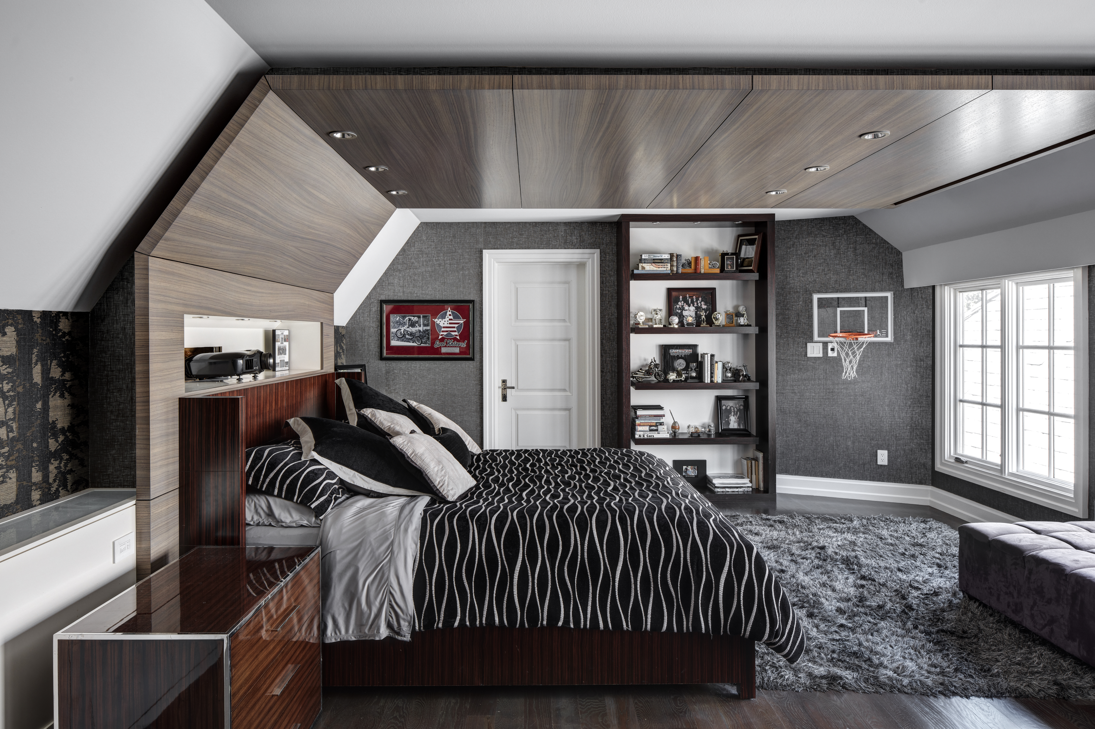 This is a stylish bedroom featuring a dark wood bed frame with a black and white patterned comforter and decorative pillows. The room has a modern aesthetic with a built-in bookshelf, a basketball hoop, and a gray textured wall. The flooring is dark wood, and a plush gray rug adds warmth to the space.