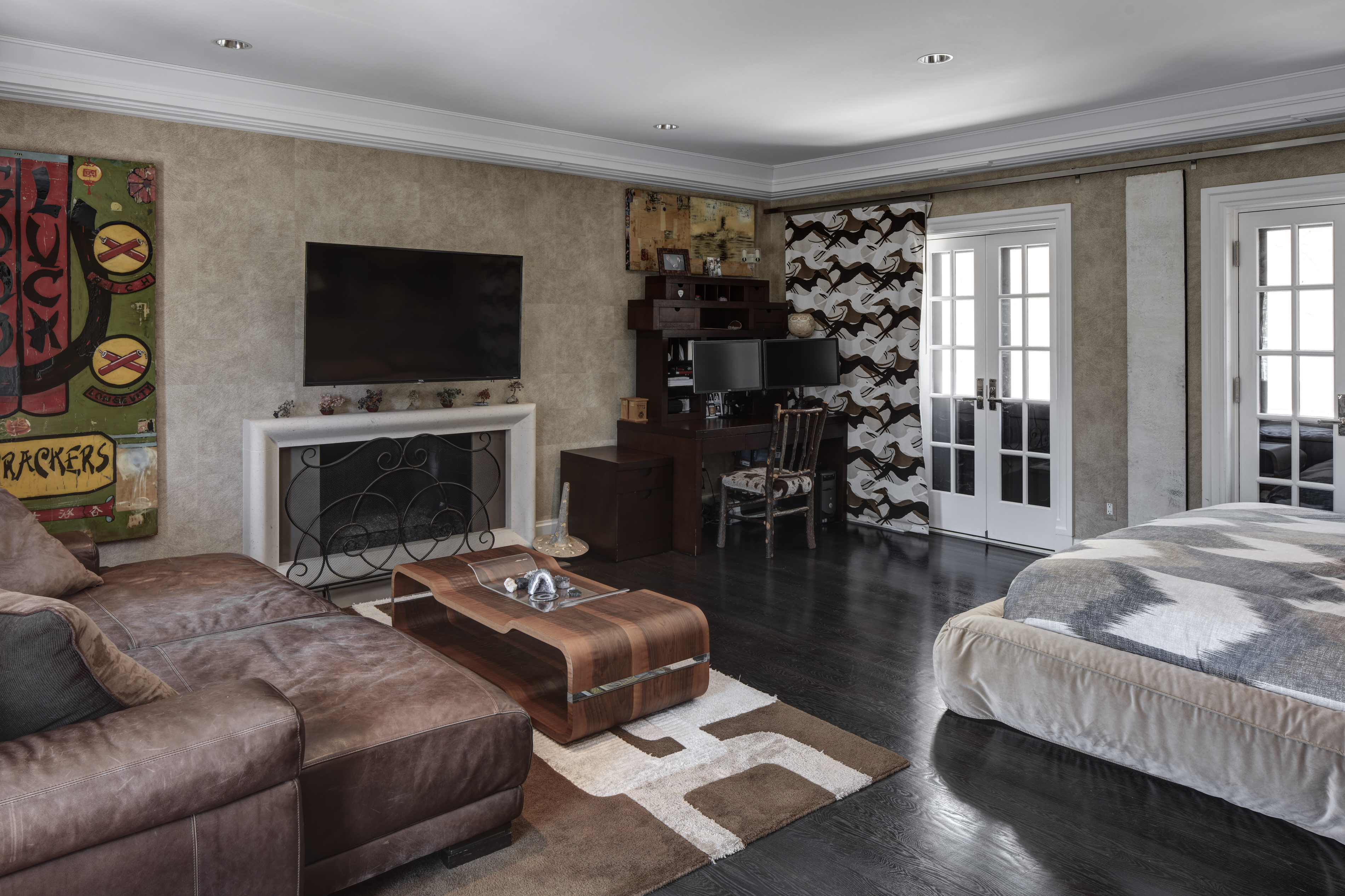 This is a primary bedroom featuring a seating area with a brown leather couch, a fireplace with a TV mounted above, and a desk area with dual monitors. The room has dark wood floors, neutral-toned walls, and white trim, creating a sophisticated and comfortable atmosphere. French doors lead to an outdoor space, and a large bed is visible on the right side of the image.