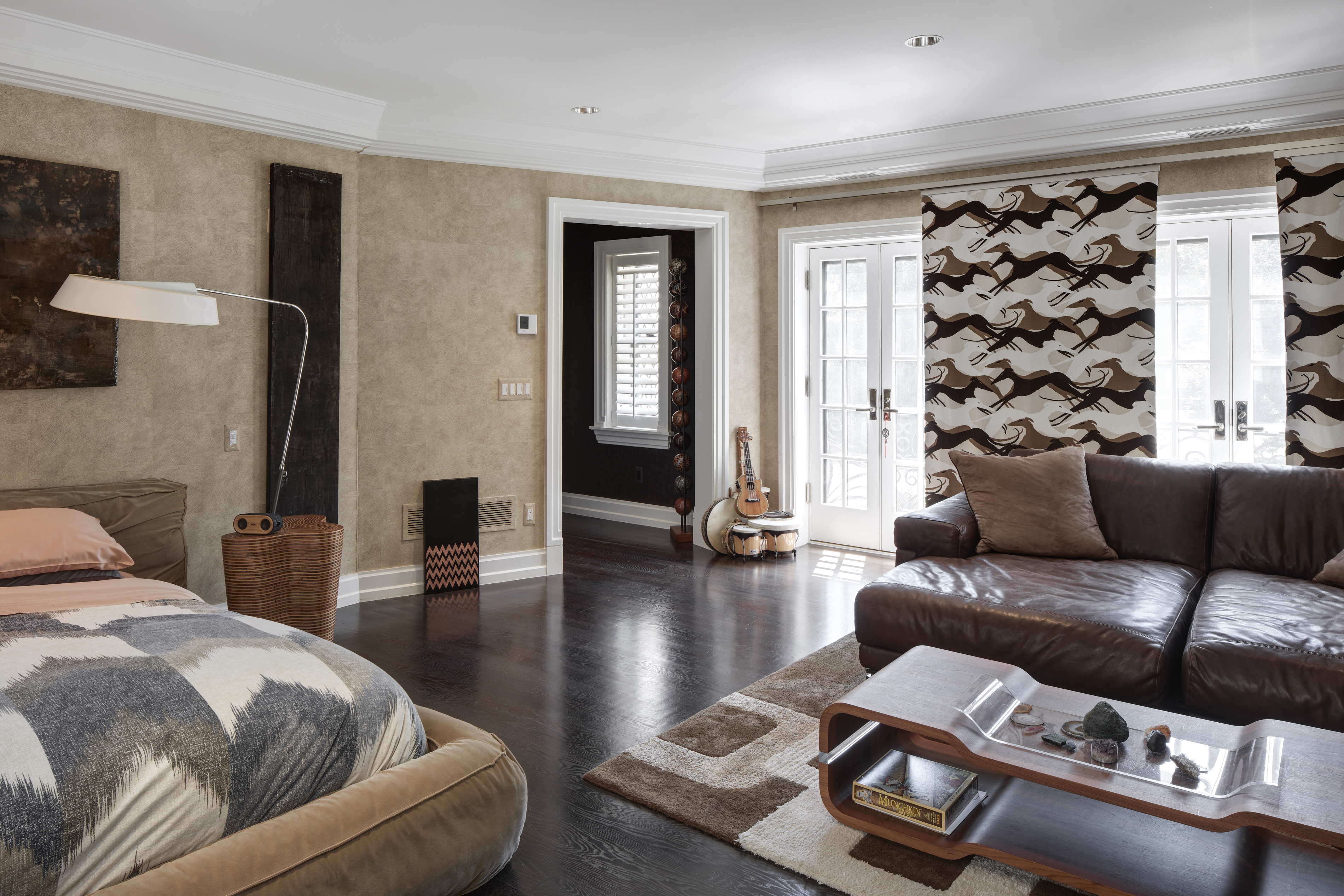 This primary bedroom features a luxurious bed with a unique patterned duvet and a plush headboard. The room is decorated with neutral tones and accented by dark wood flooring and modern art pieces. French doors lead to an outdoor area, enhancing the room's spacious and airy feel.