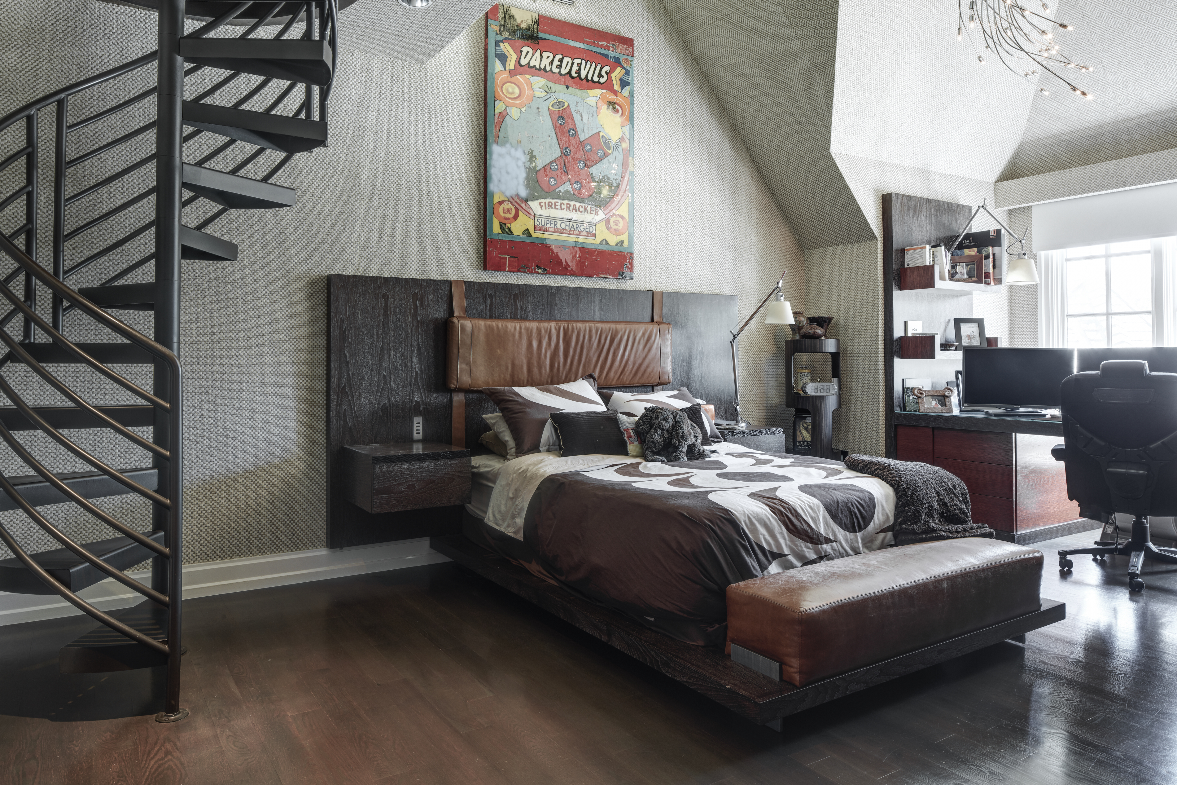 This is a stylish primary bedroom featuring a unique spiral staircase and a modern bed with a dark wood frame and leather headboard accents. The room includes a dedicated workspace with a desk and chair, complemented by a vintage-inspired 'Daredevils' poster above the bed. The perspective is from the foot of the stairs, showcasing the room's layout and design elements.