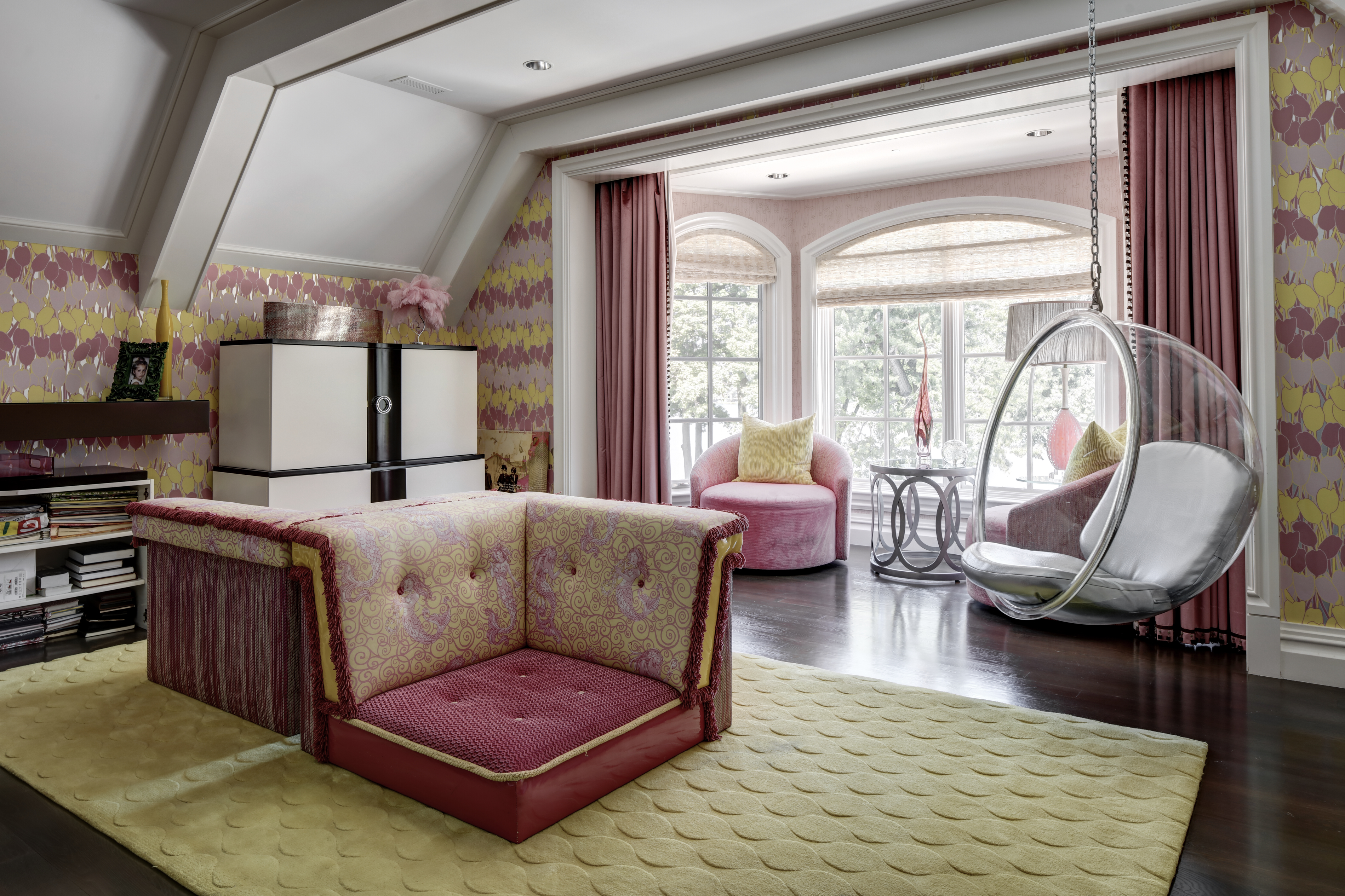 This interior shot showcases a stylish living room with unique design elements. The room features a custom-designed corner sofa, a transparent hanging bubble chair, and a pink accent chair, all set against a backdrop of patterned wallpaper and dark hardwood floors. The space is bright and inviting, with a playful yet sophisticated aesthetic.