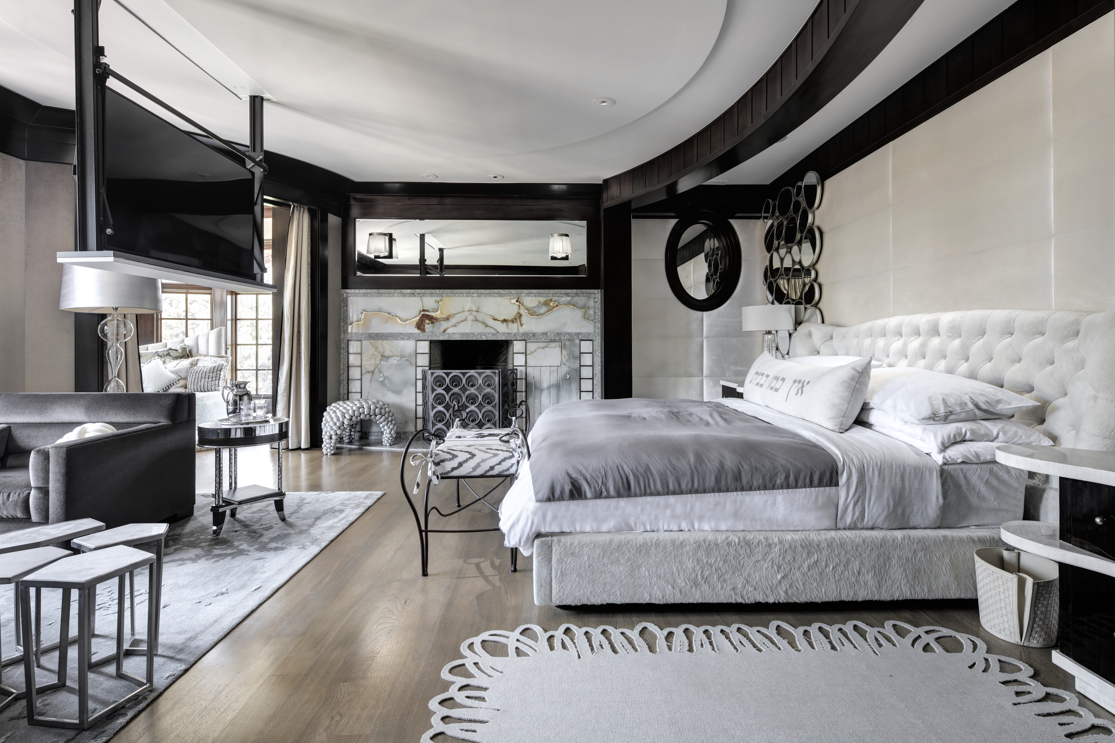 This is a luxurious primary bedroom featuring a large bed with a tufted headboard, a fireplace with a marble surround, and a seating area with a dark gray sofa. The room has a sophisticated and modern design with dark wood accents and a light-colored rug. A large television is mounted on a ceiling fixture.