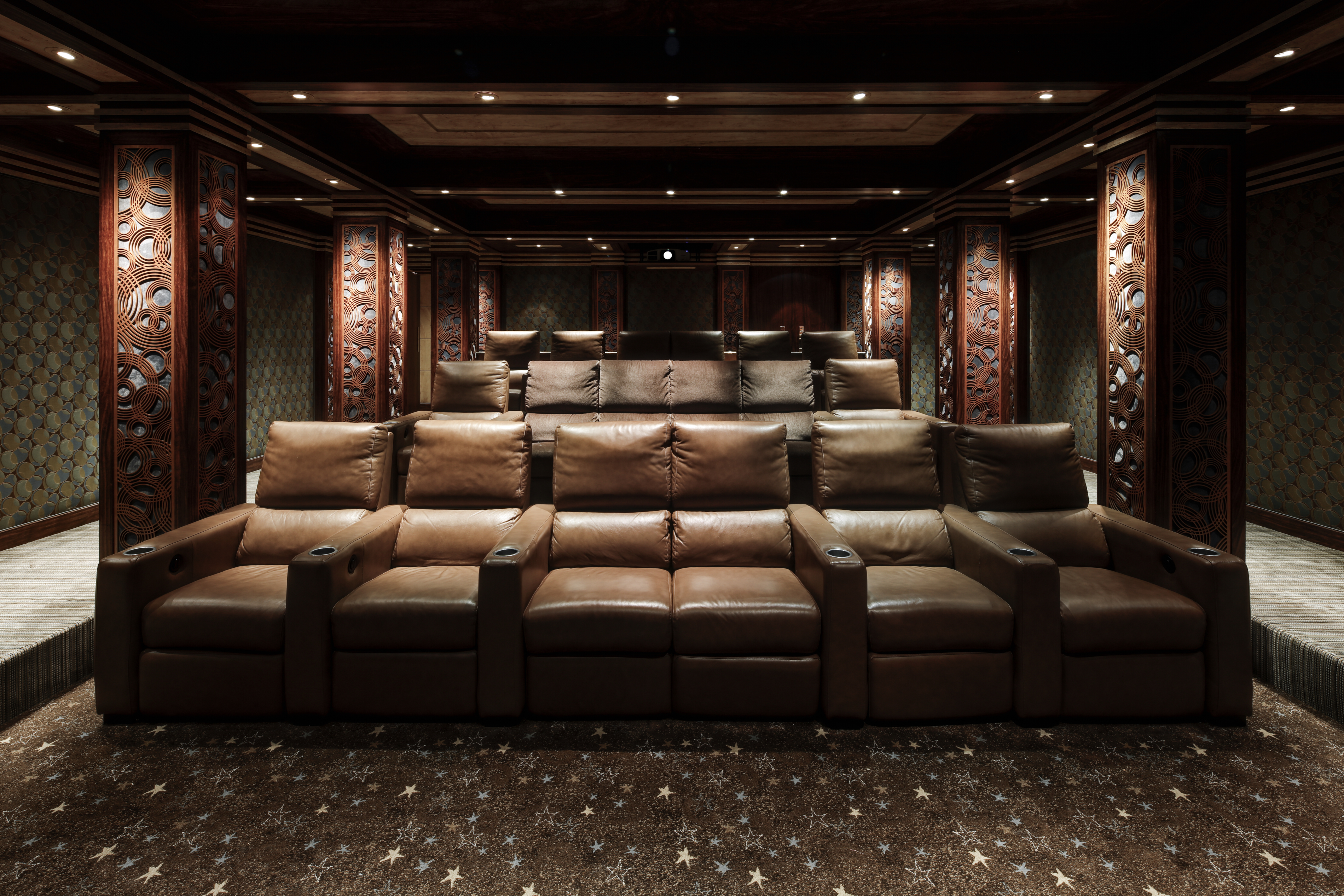 This is an interior shot of a luxurious home theater. The room features multiple rows of plush, brown leather reclining seats with cup holders, arranged on tiered platforms. The walls are adorned with decorative wood paneling and patterned wallpaper, while the ceiling has recessed lighting, creating a sophisticated and immersive viewing experience.