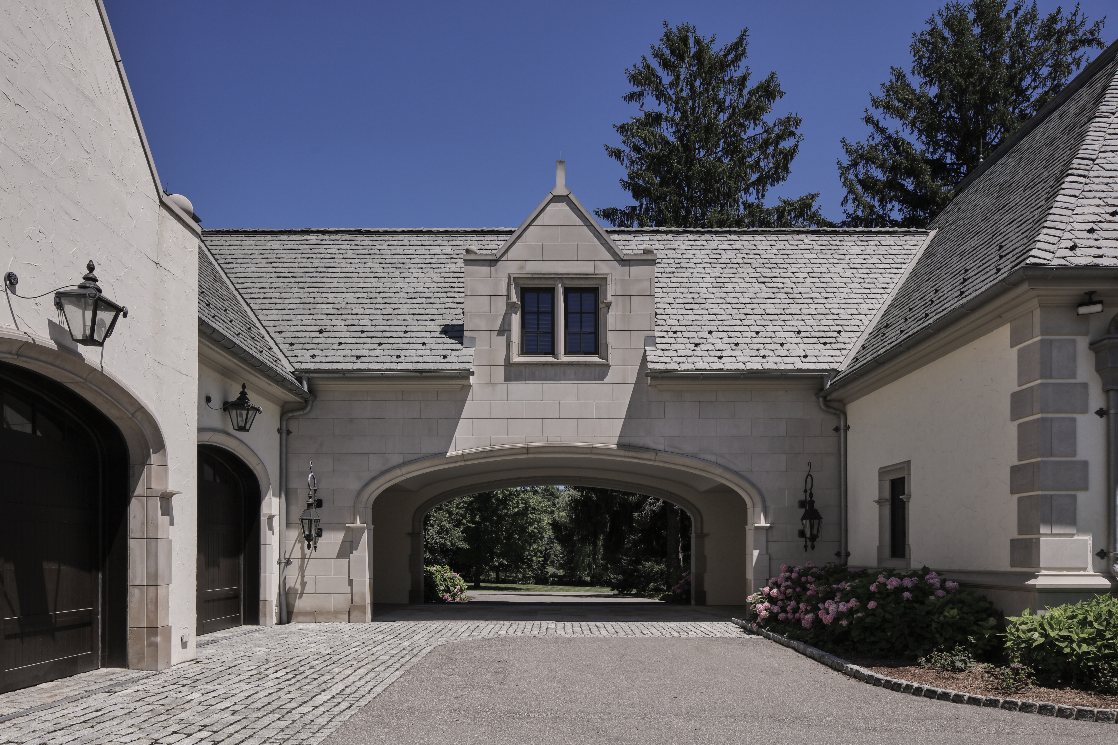 This image showcases a grand entryway to a property, featuring a stone archway connecting two wings of the building. The structure is adorned with elegant exterior lighting fixtures and the driveway is paved with a combination of cobblestone and asphalt. Lush landscaping, including pink hydrangeas, adds a touch of color and sophistication to the scene, creating a welcoming and impressive first impression.