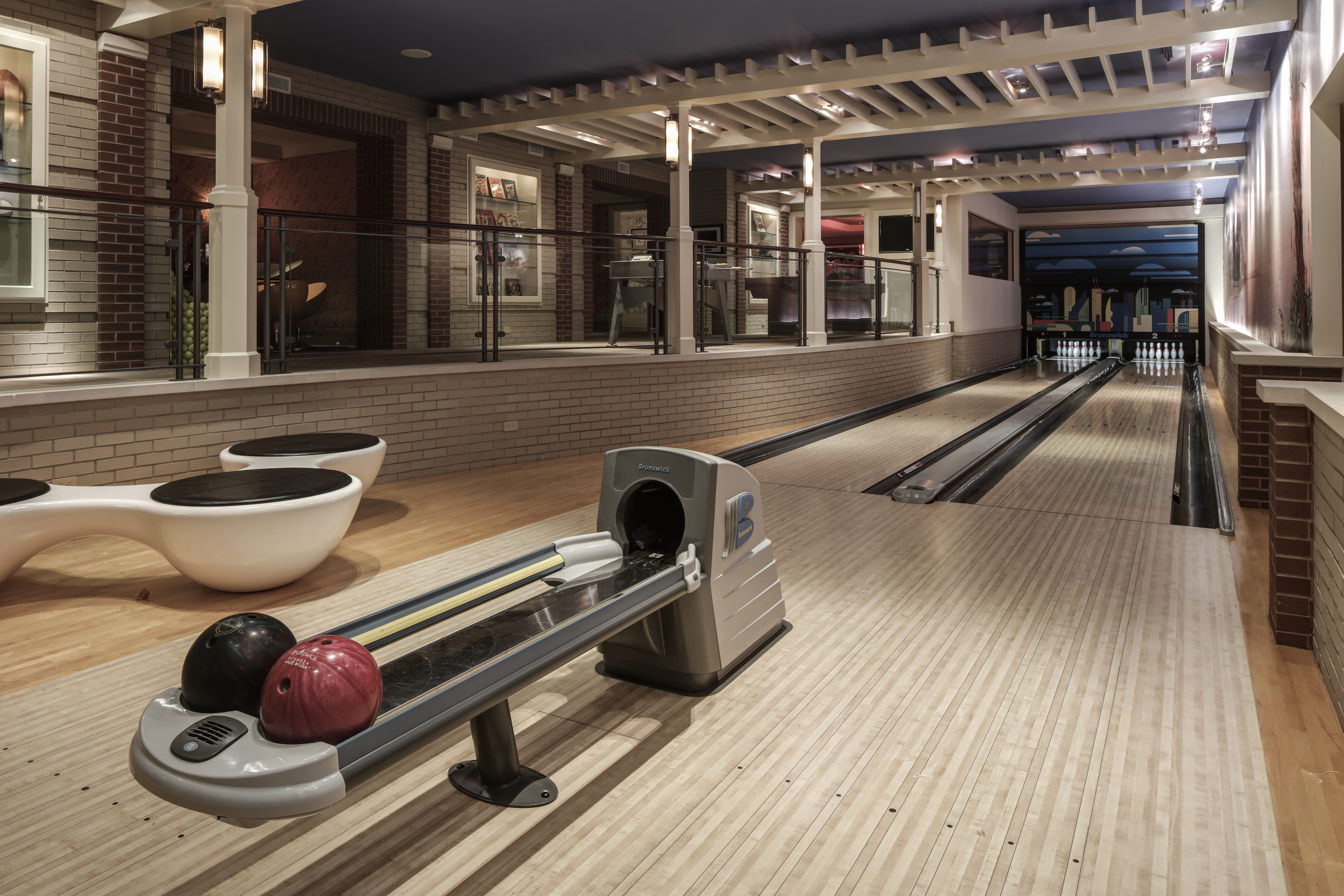 This interior shot showcases a well-appointed bowling alley within a home. The lanes are pristine, flanked by comfortable seating and modern bowling equipment. The space features a sophisticated design with brick accents and a stylish overhead structure, creating a luxurious recreational area.