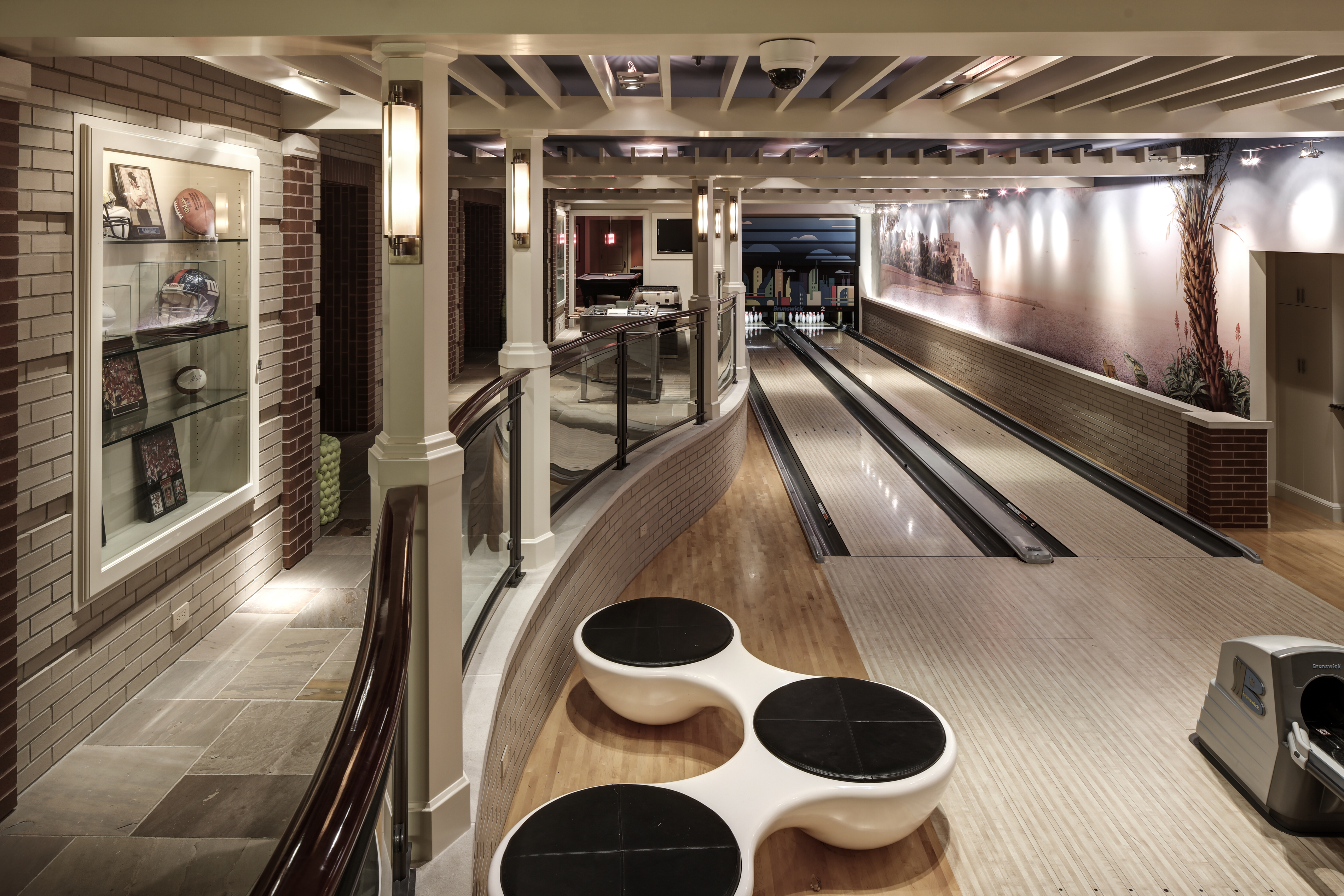 This is a spacious basement featuring a custom bowling alley with two lanes, a seating area with unique circular benches, and a display case showcasing sports memorabilia. The design incorporates brick accents, wood flooring, and a coffered ceiling with recessed lighting, creating a luxurious and entertaining space. The perspective is from the entrance of the bowling alley.